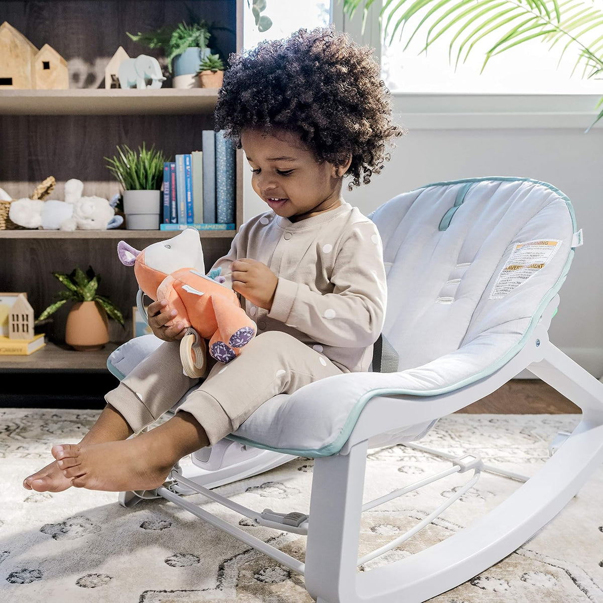 Toddler seated in a light-gray bouncer on a rug, smiling and holding a plush toy.