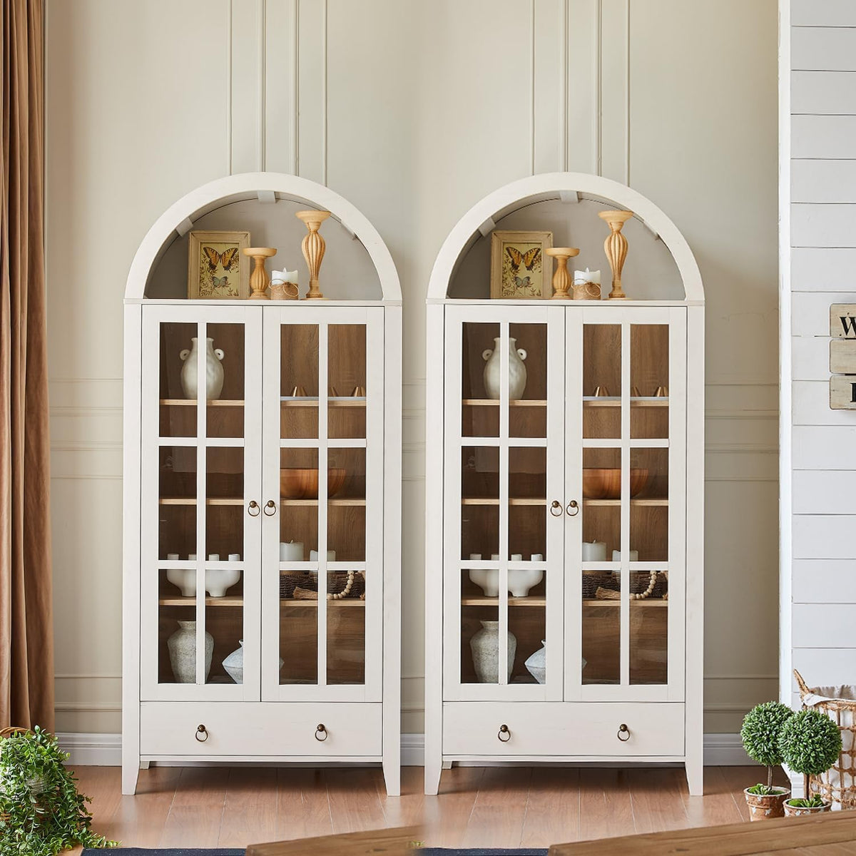 Two white-finish arched cabinets staged together with warm wood interiors and tabletop decor above