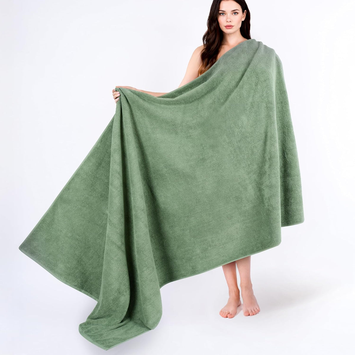 Woman holding a large sage-green towel wrapped across her body, standing barefoot on a white background.