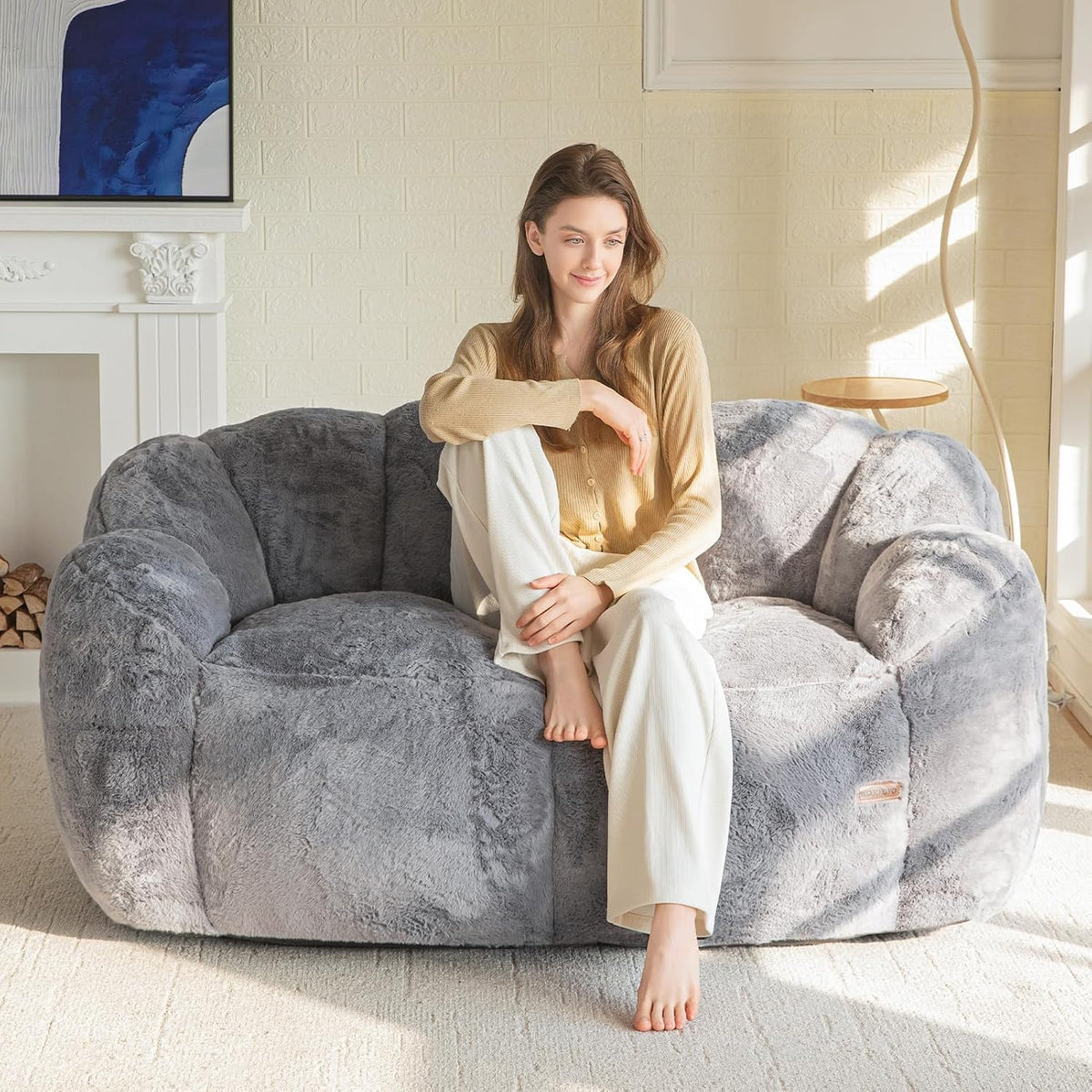 Woman perched on a large plush gray floral-shaped bean bag sofa by a fireplace and floor lamp.