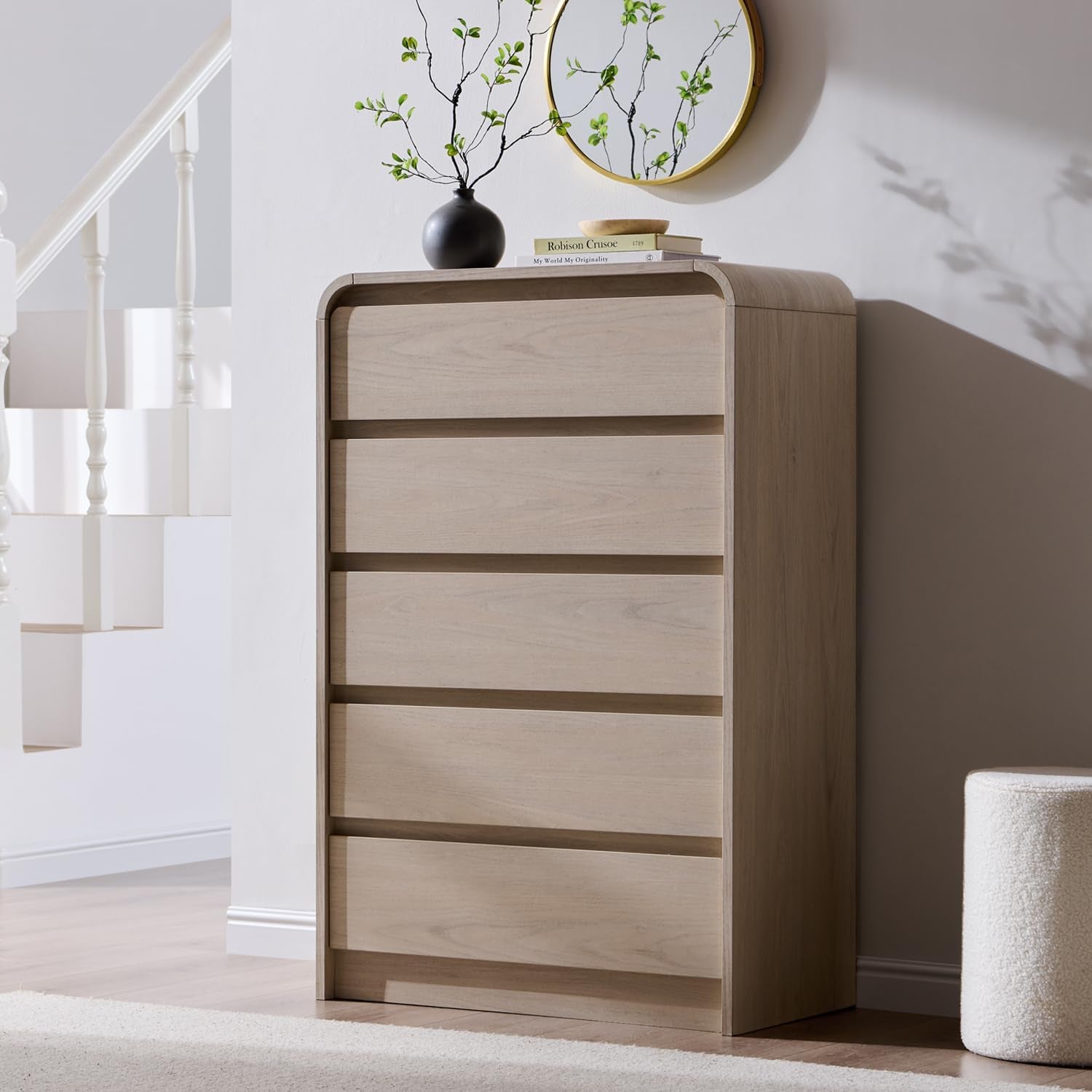 Oak tall dresser in a bright entryway next to a white staircase, topped with a black vase and round mirror.