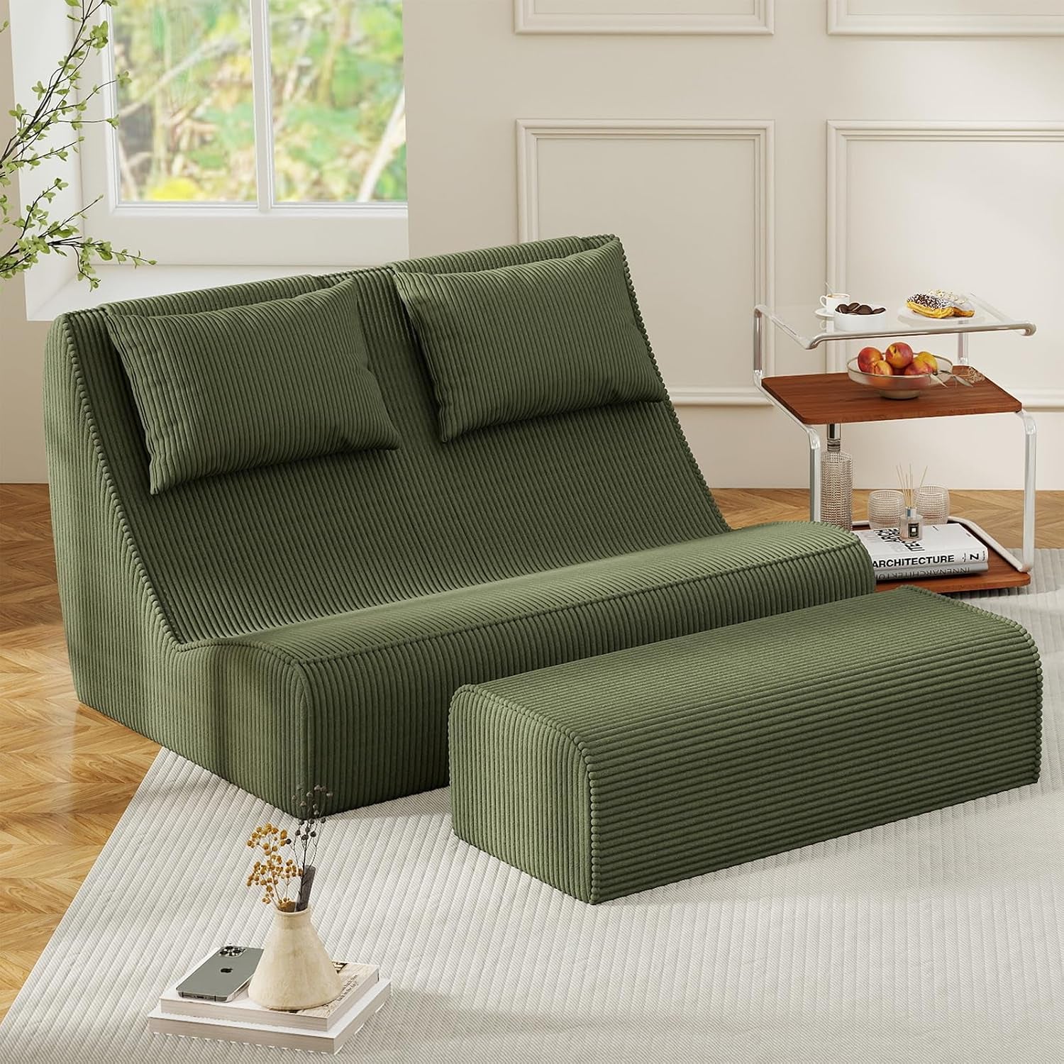 Olive-green two-seat chaise with two back cushions and matching ottoman, paired with a clear side table topped with fruit.