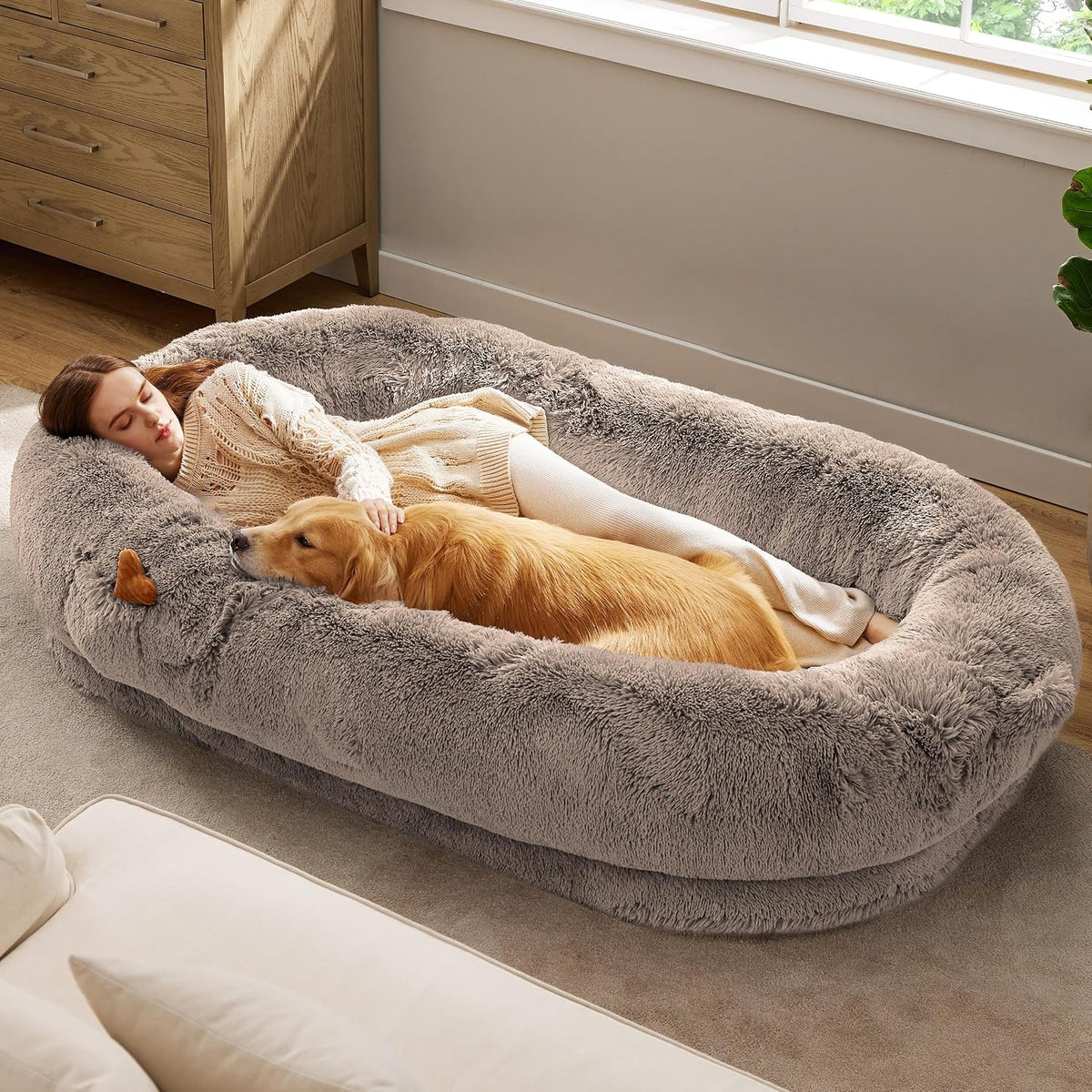 Large gray faux-fur donut bed on the floor with a woman and a golden retriever lying inside