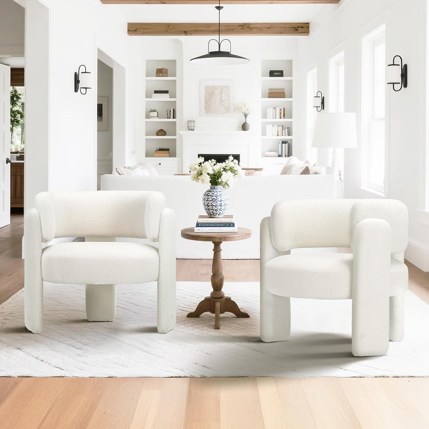 Pair of white rounded barrel chairs with three-legged block bases flanking a small wooden side table in a bright living room.