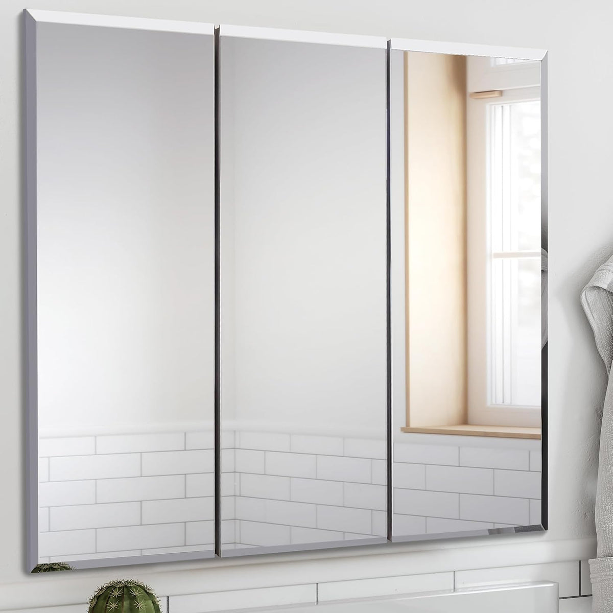 Wall-mounted three-panel mirrored medicine cabinet above a white tiled backsplash, reflecting a window and towel