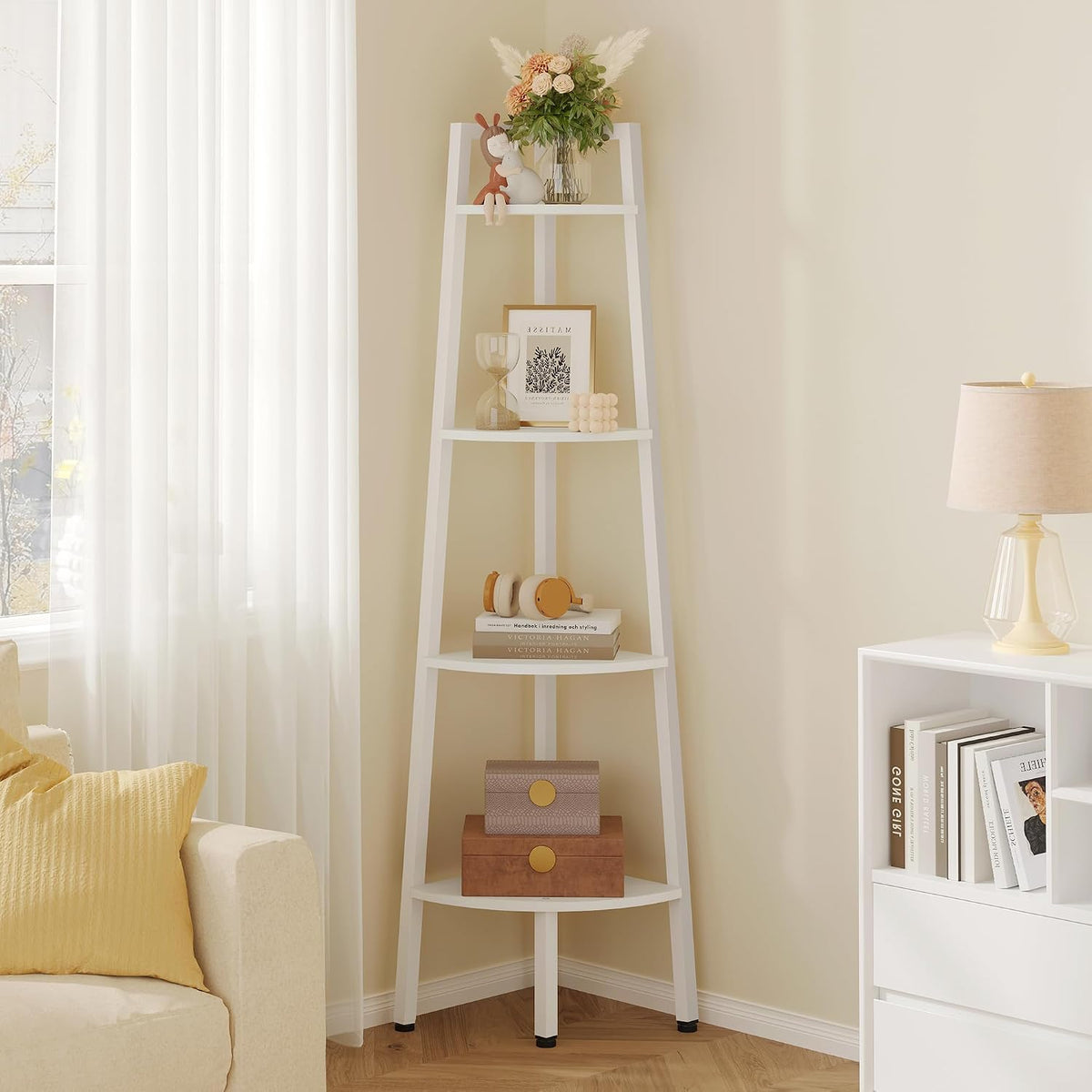 White corner ladder shelf styled beside a sheer-curtained window and tabletop lamp