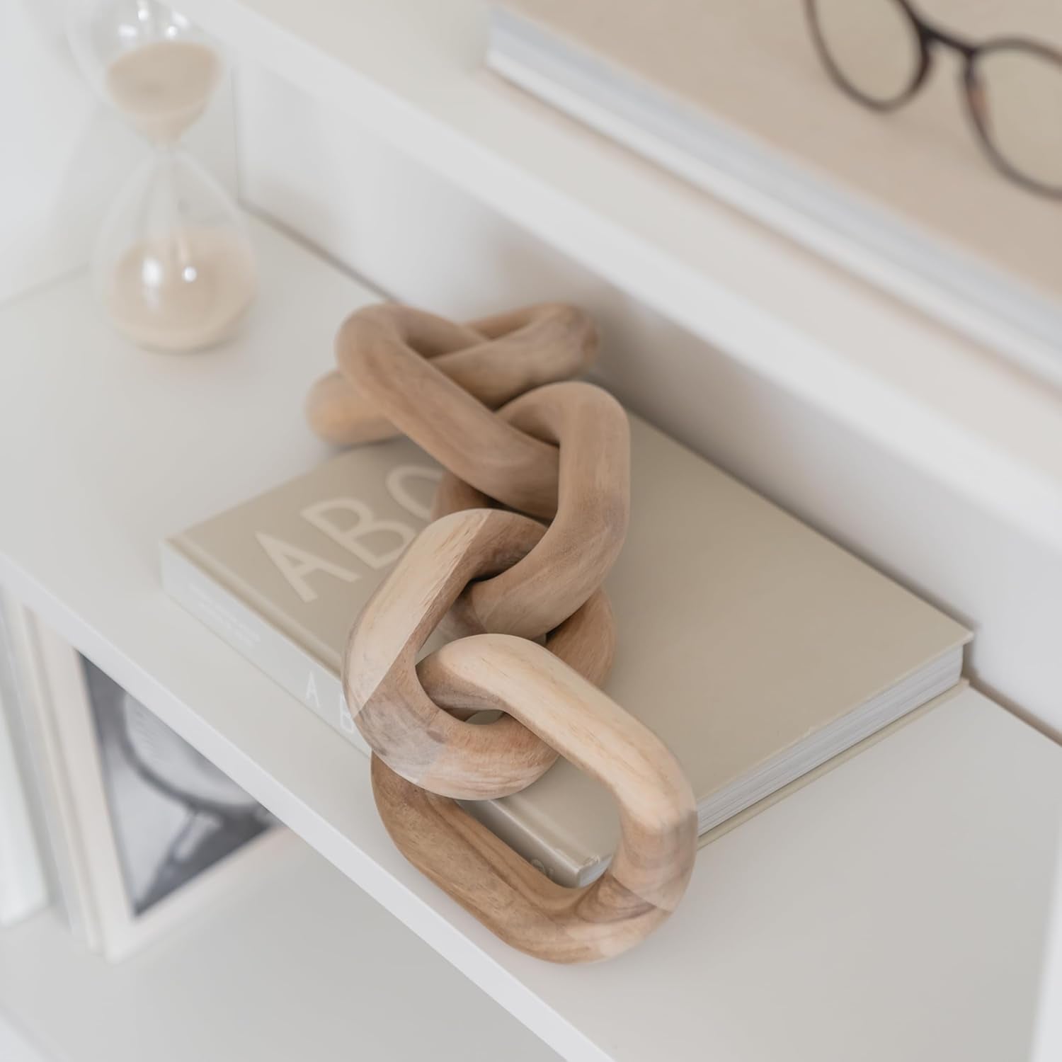 Carved acacia link chain coiled on a beige book on a white shelf, soft top-down view with hourglass nearby.