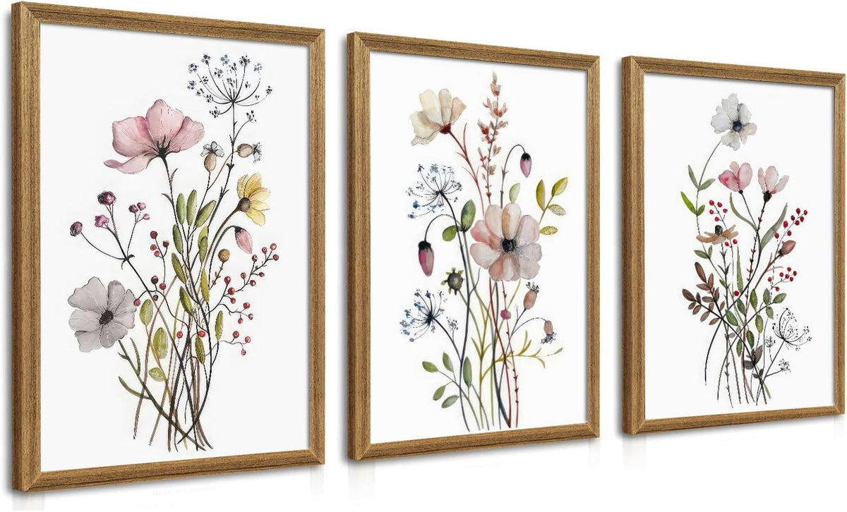 Angled view of three vertical framed botanical prints with soft pink and muted tones in natural wood frames on a white