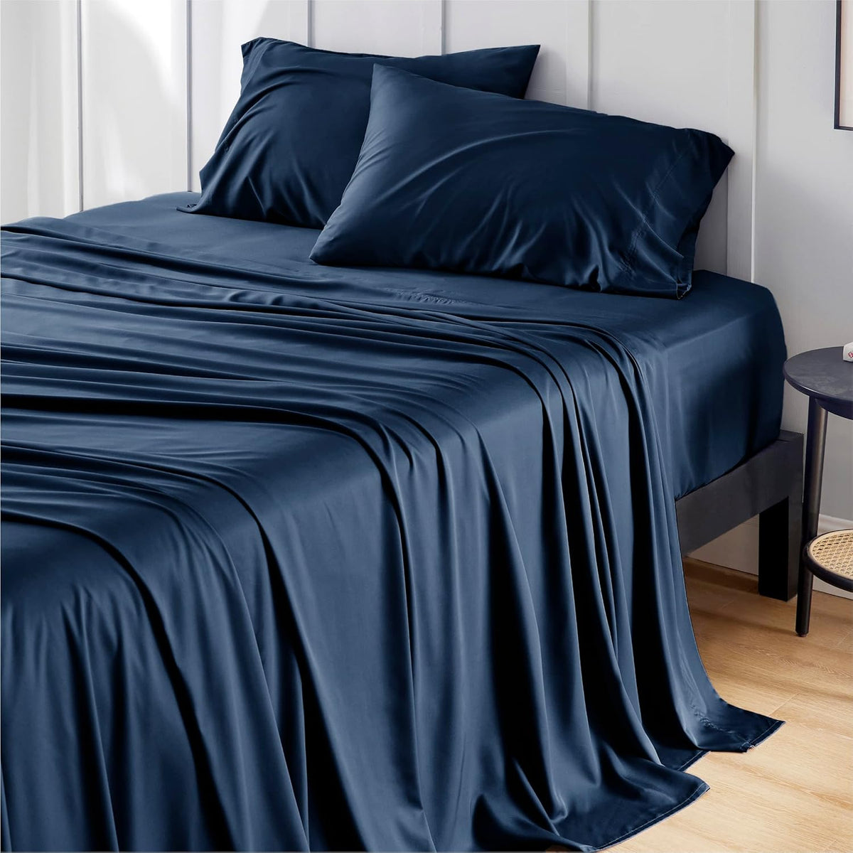 Deep navy silky sheets and two matching pillows arranged on a bed with a white paneled wall.
