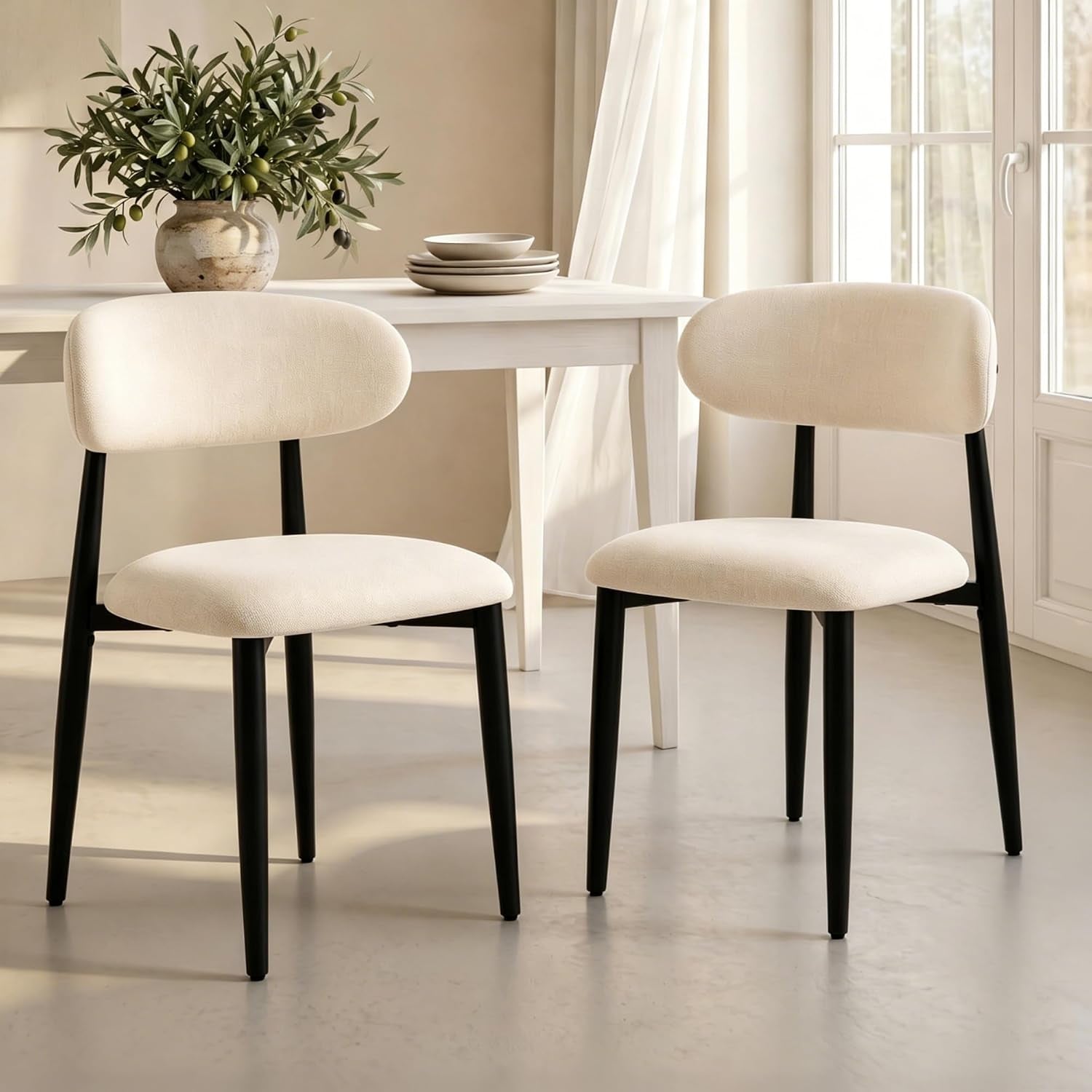 Two beige-upholstered dining chairs with rounded backs and matte black metal legs beside a white table by a window.