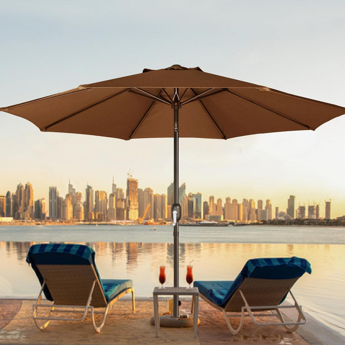 Brown umbrella shading two lounge chairs beside calm water with a city skyline in the background.