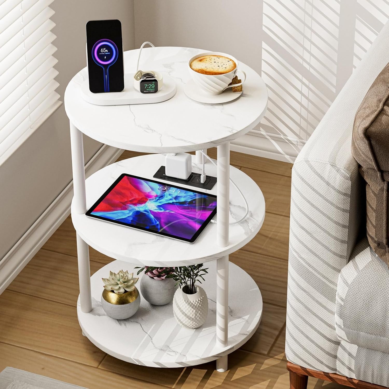 Round three-tier white marble-look side table beside a sofa with phone on a wireless charger, tablet, and small planters