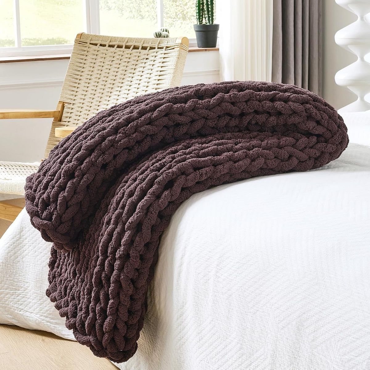 Deep brown chunky knit throw rolled and piled at the bed edge with a small cactus on the windowsill behind.