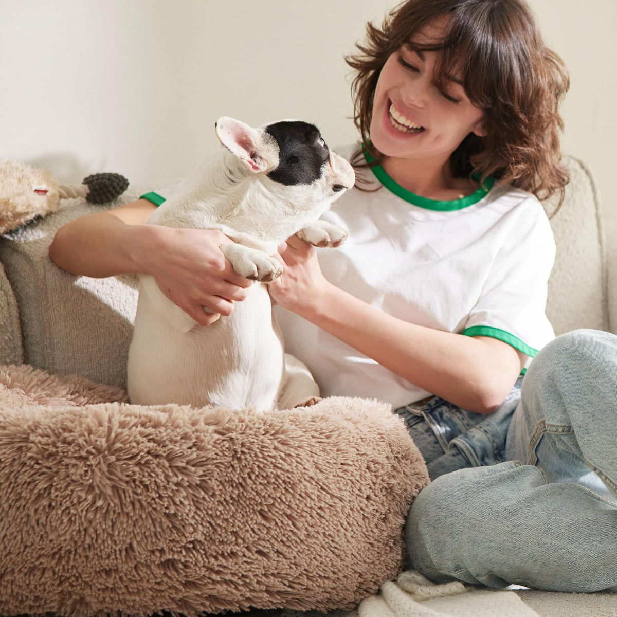 Woman on couch holding a small black-and-white dog sitting beside a tan fluffy round dog bed