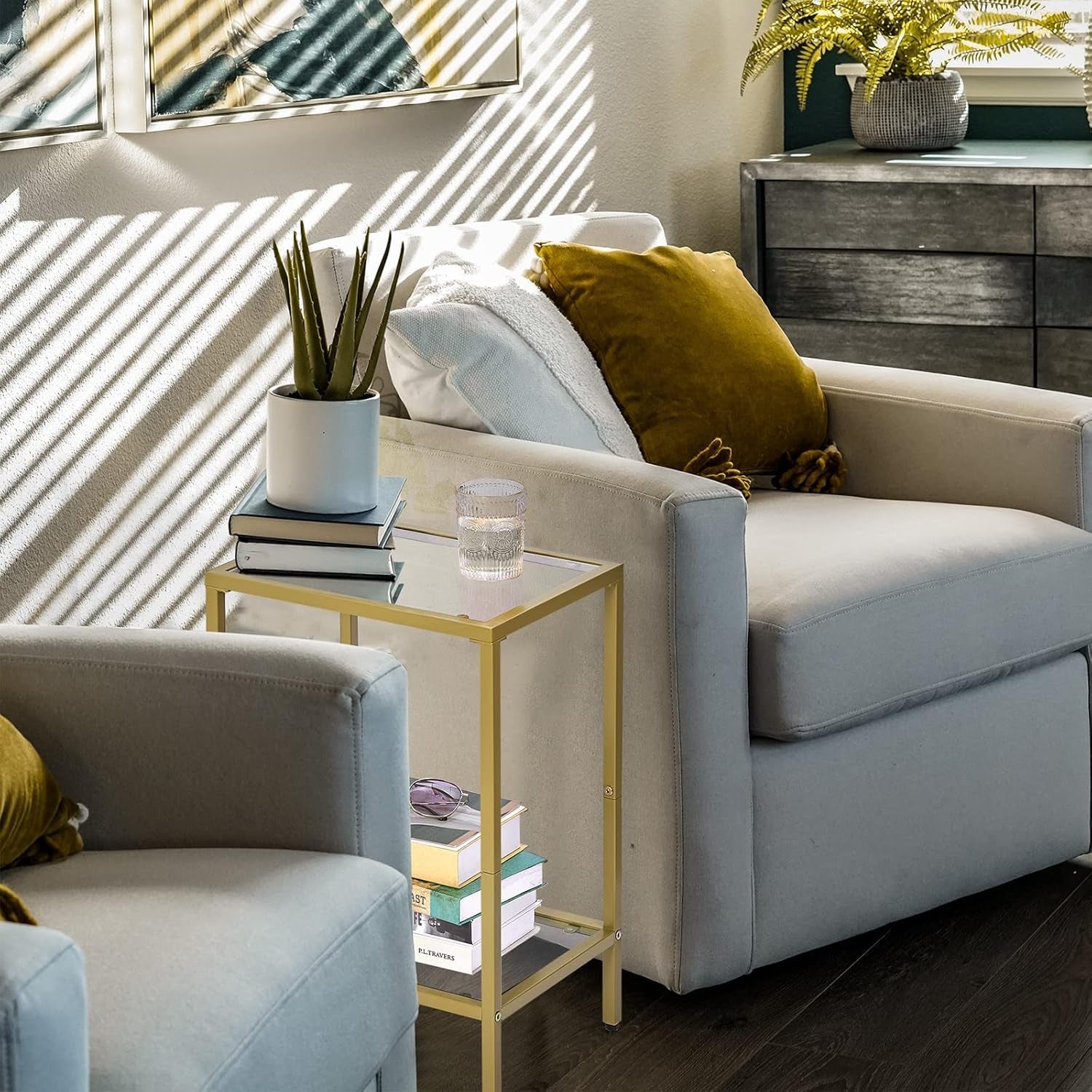 Gold side table between two gray armchairs holding a plant, books and a glass, sunlight casting striped shadows