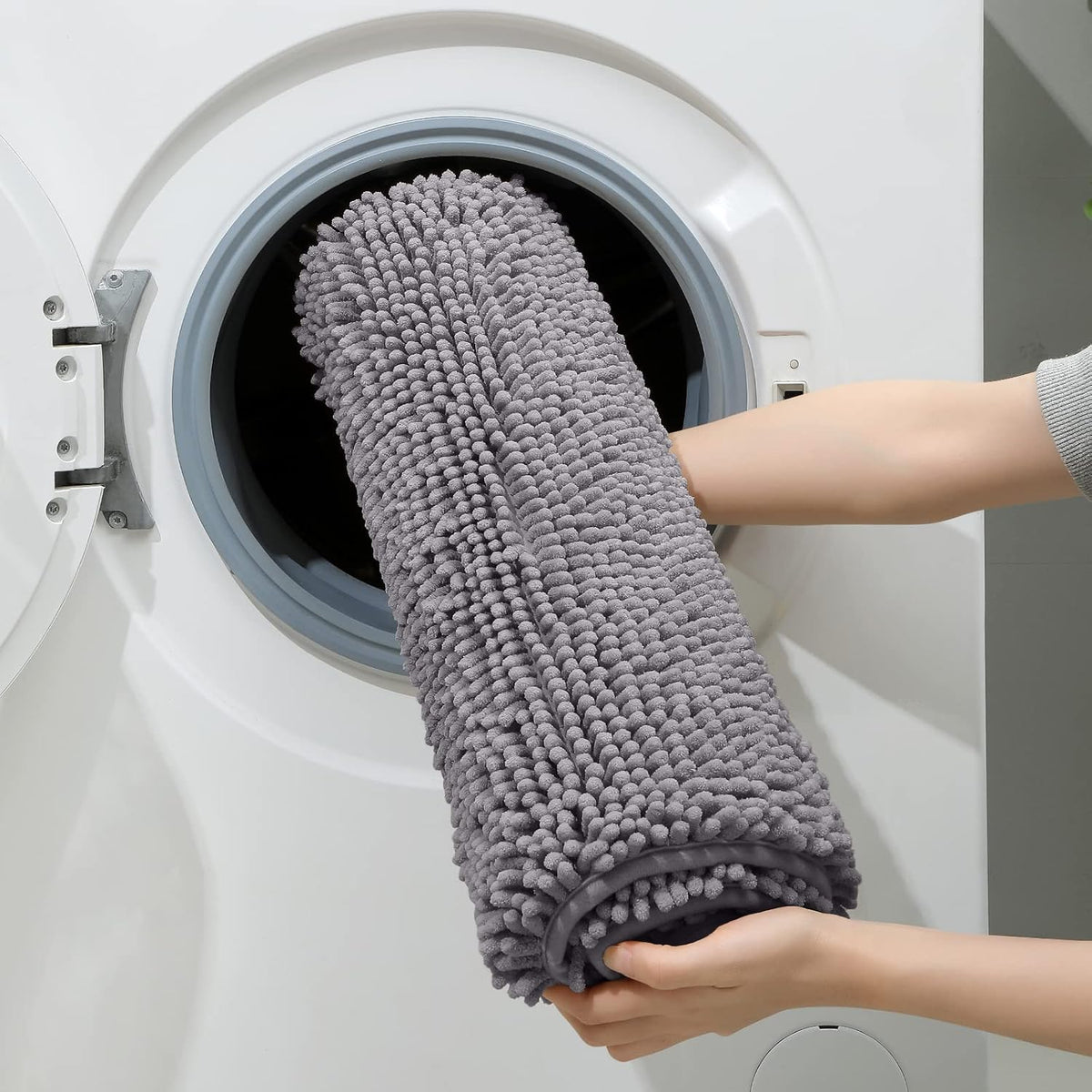 Two hands loading a rolled grey chenille bath mat into a front‑load washing machine.