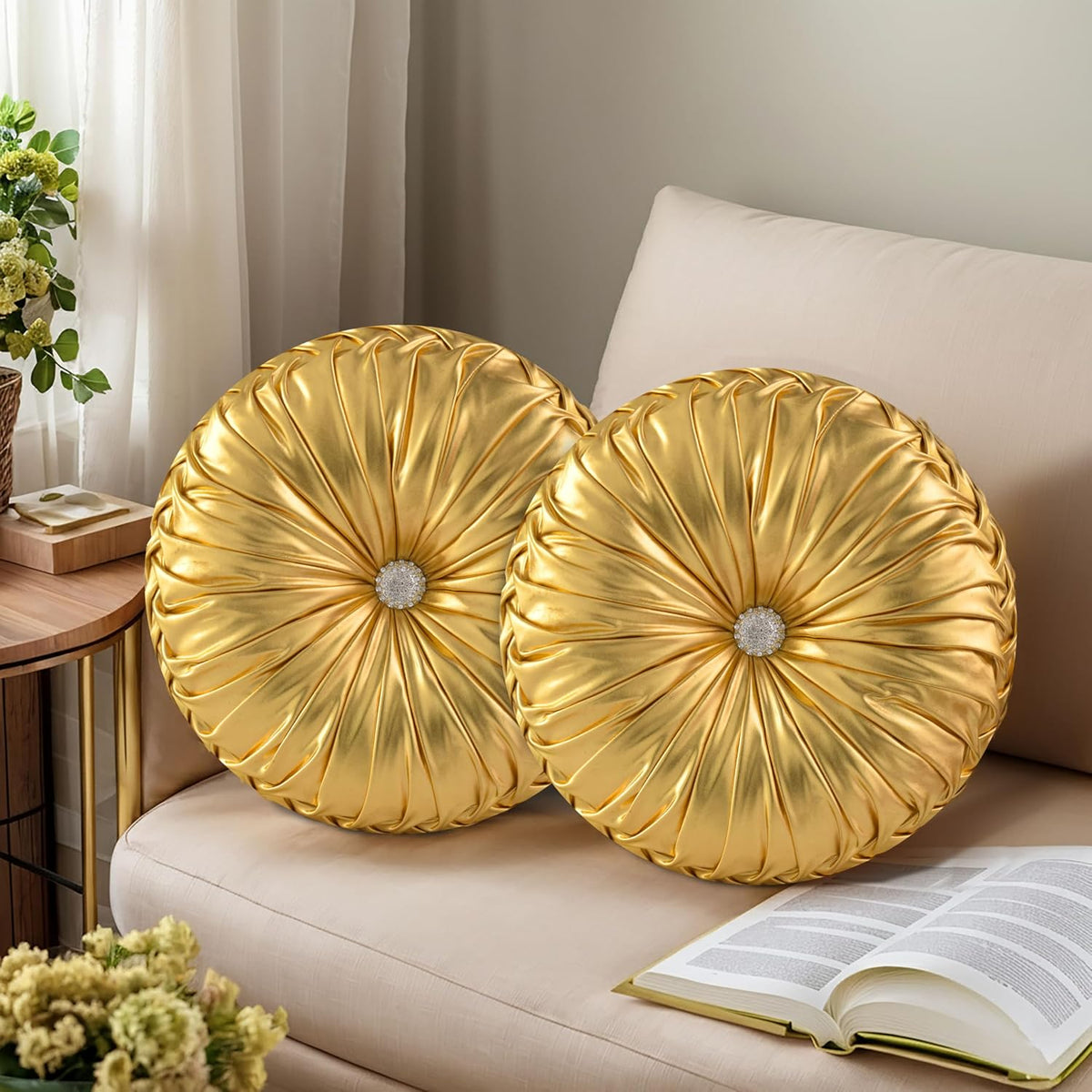 Pair of round gold ruched pillows with sparkly centers stacked on a beige couch next to an open book.
