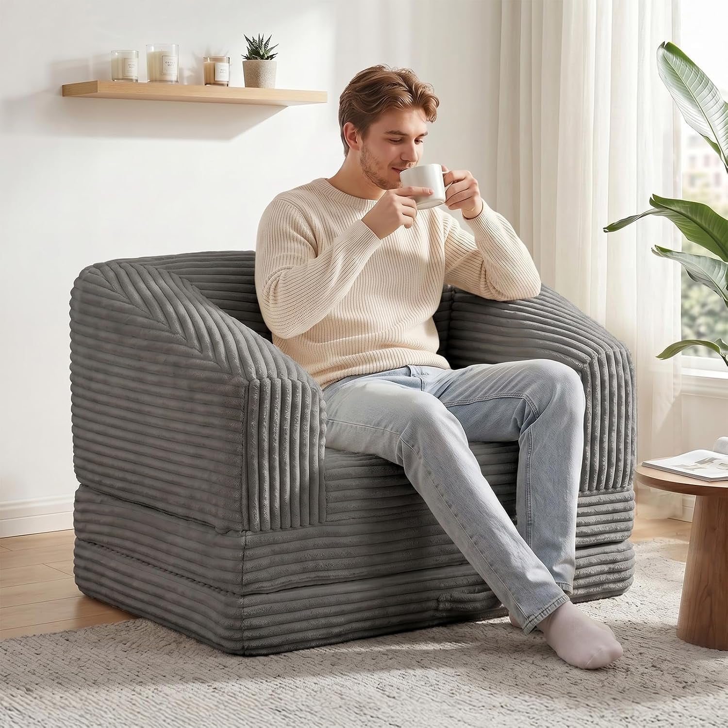 Man drinking from a mug while sitting on a dark gray corduroy folding sofa in a cozy room.