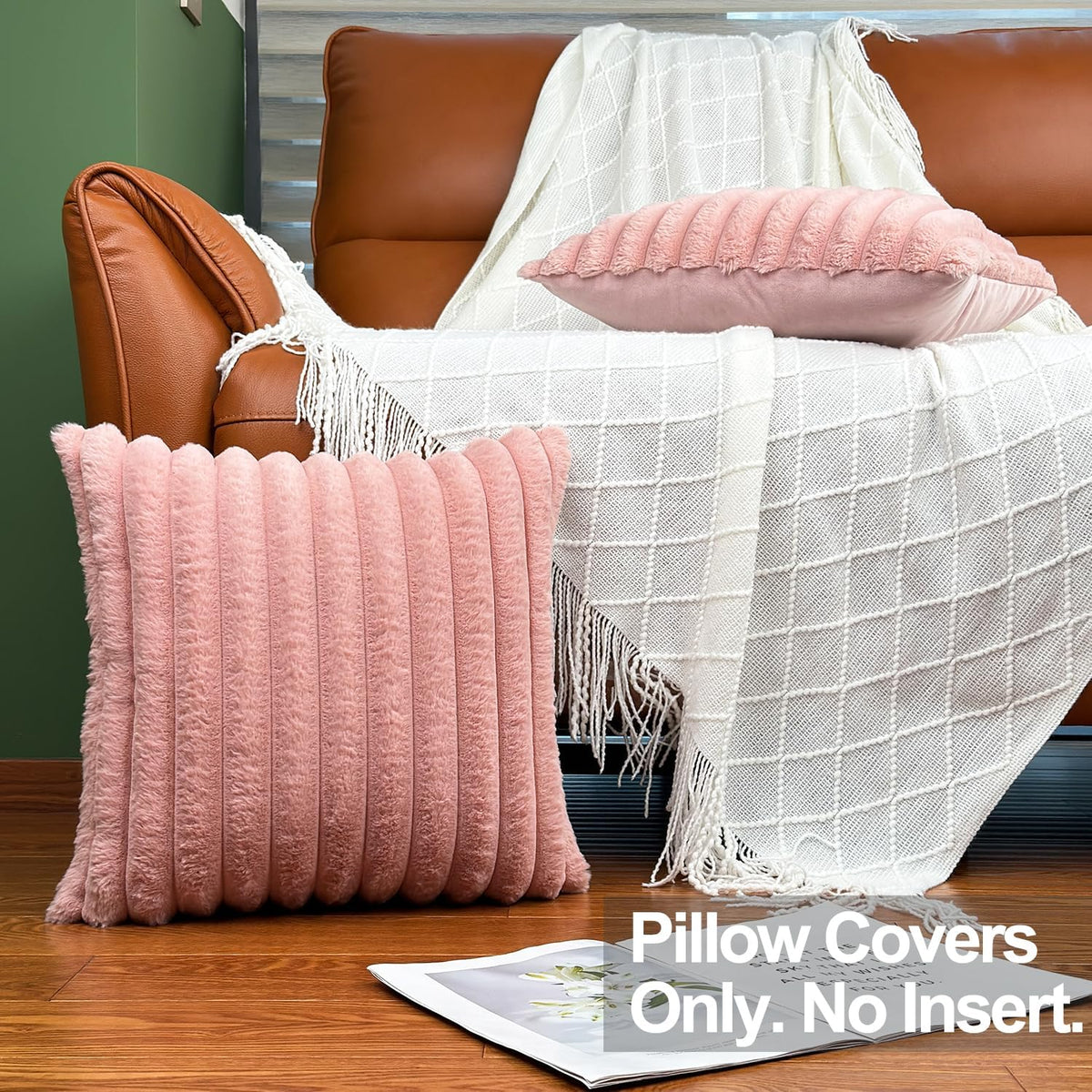 Pink ribbed faux‑fur pillow leaning on the floor beside a matching pillow resting on a white fringed throw over a brown