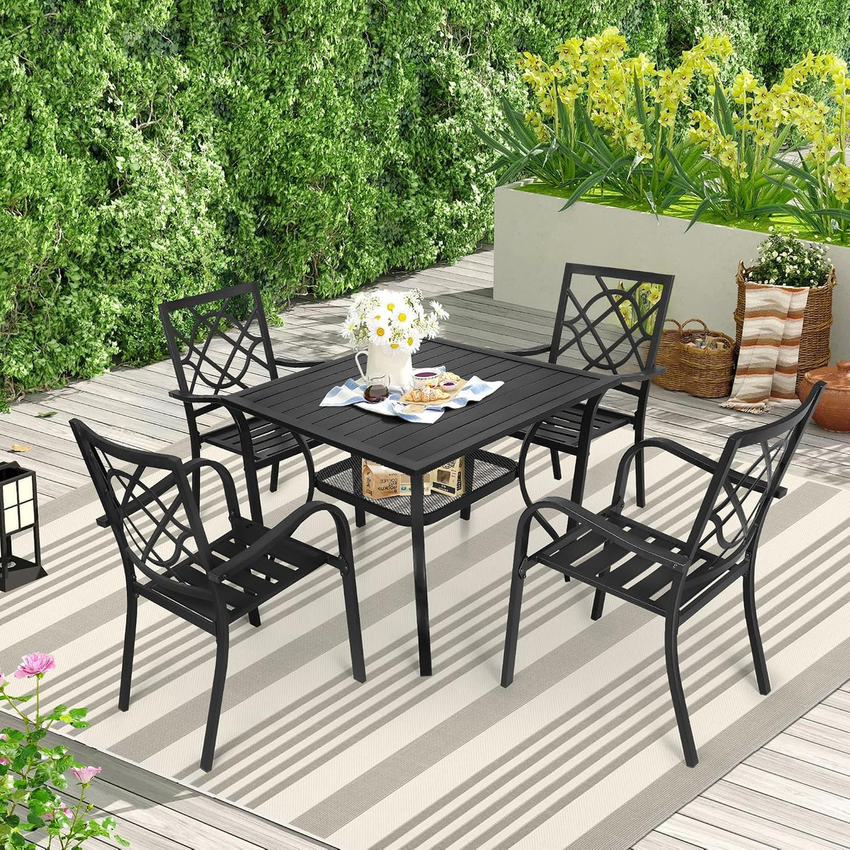 Square black metal table with four matching chairs on a striped outdoor rug beside lush greenery and planters