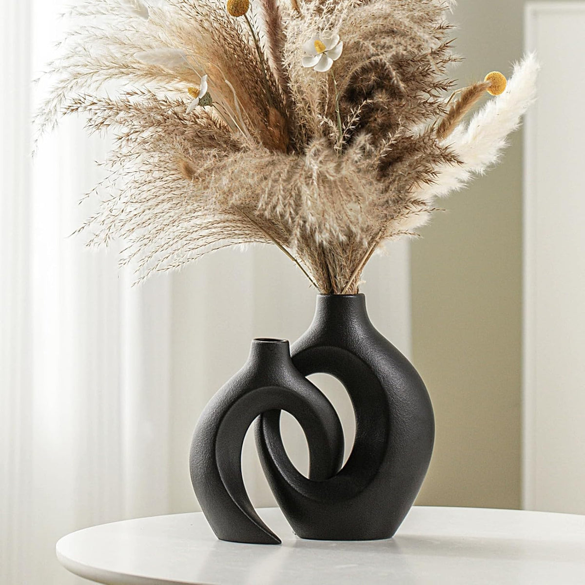 Pair of black hollow ceramic vases linked together on a white round table topped with pampas grass.