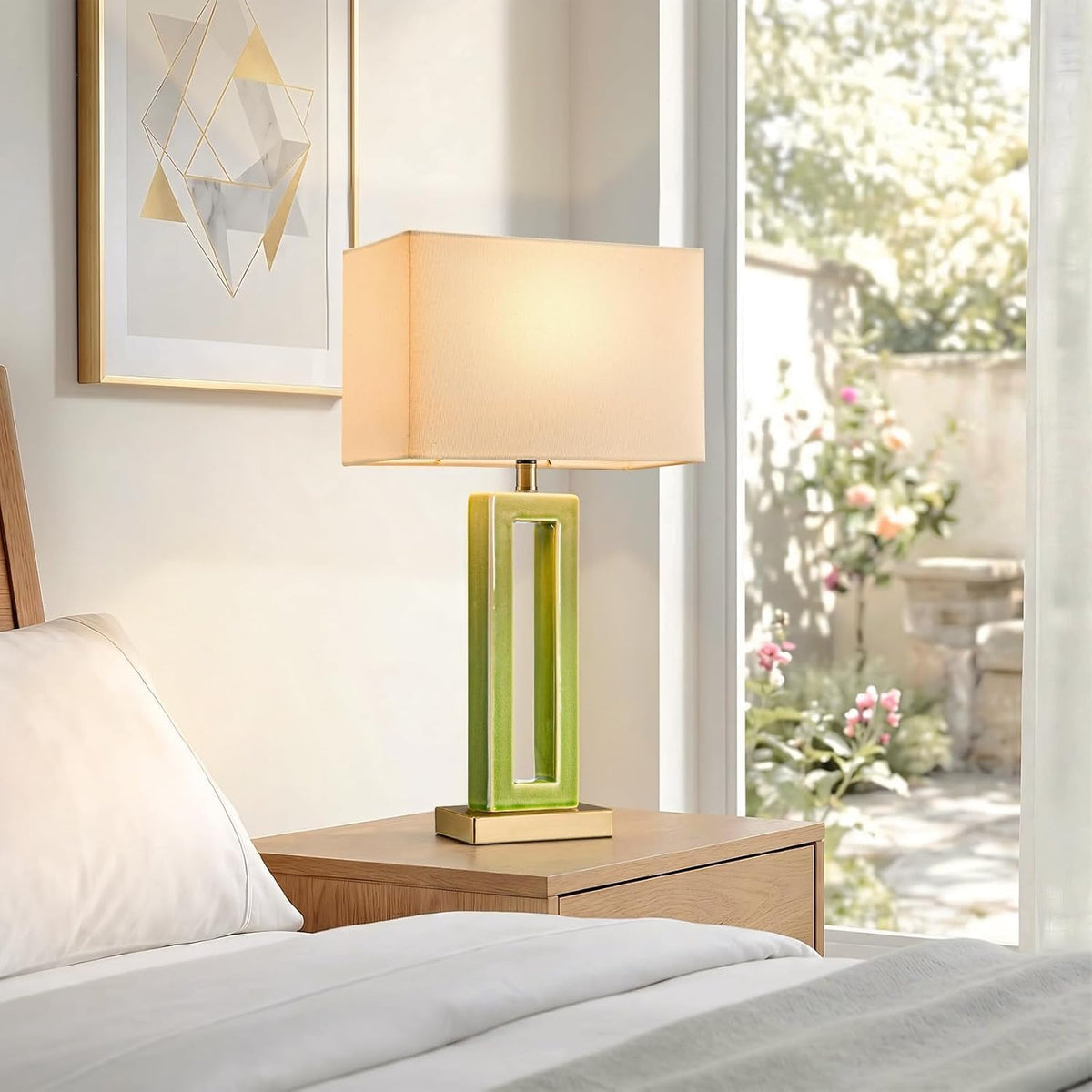 Single green hollow ceramic lamp with a rectangular shade on a wooden nightstand beside a bed and window garden view.