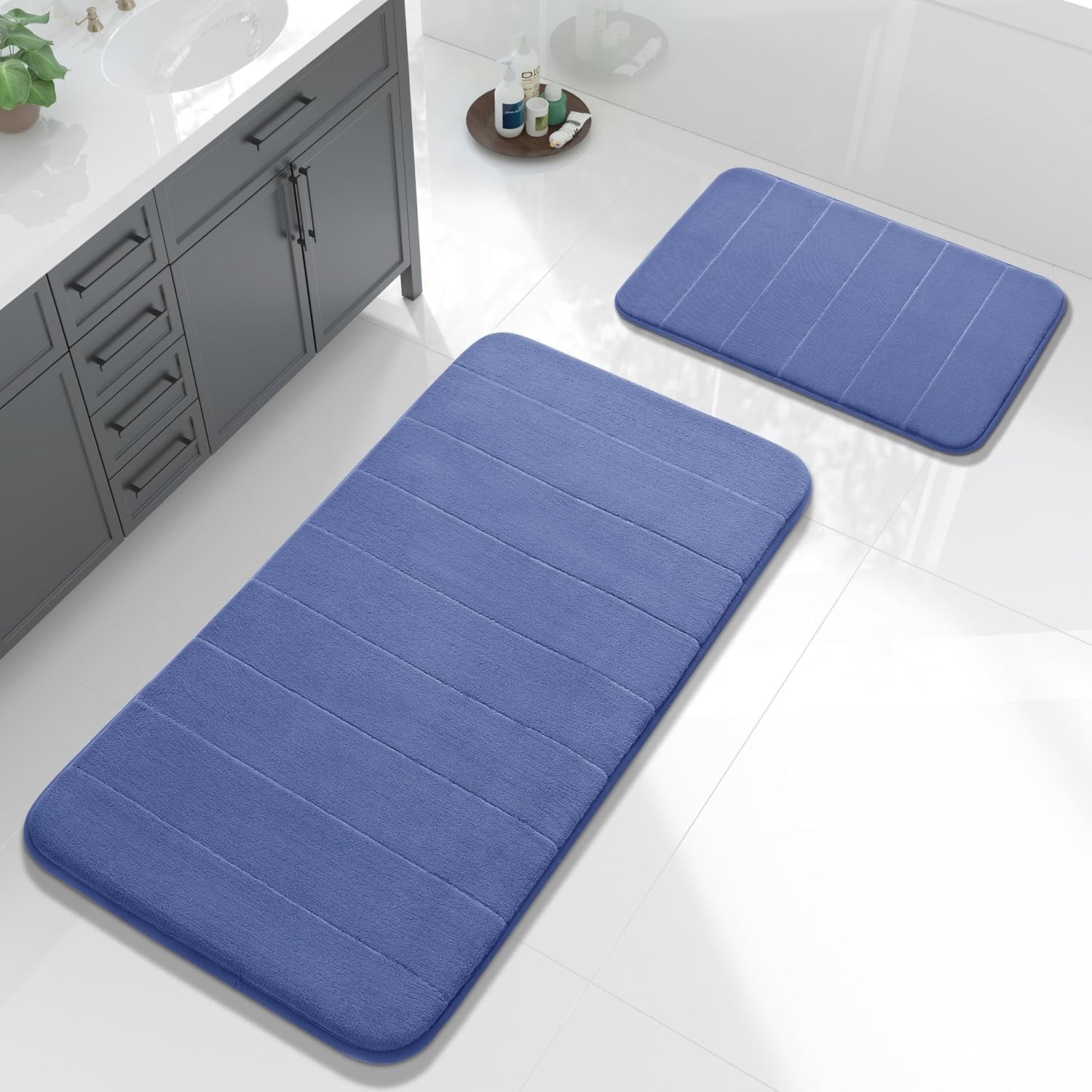 Two blue memory-foam bath mats on glossy white tile beside a gray vanity and toiletries.