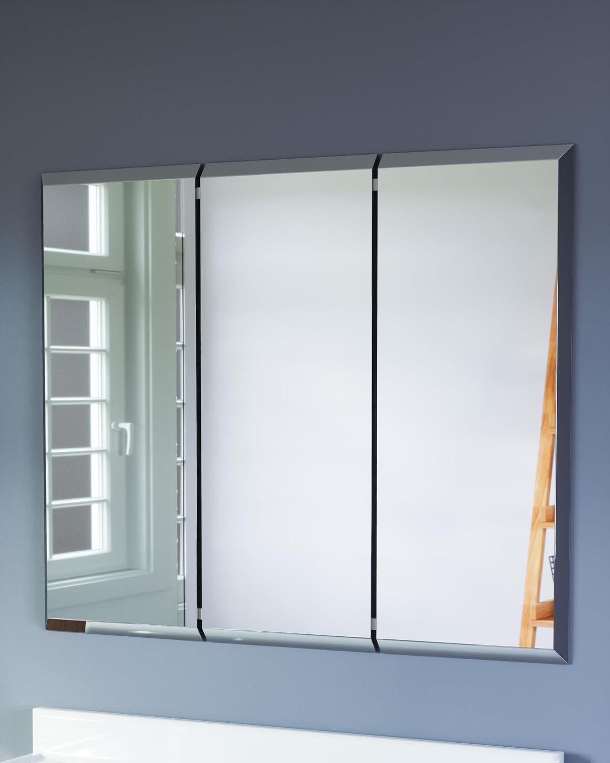 Three-panel mirrored medicine cabinet mounted on a blue wall reflecting a window and wooden ladder shelf