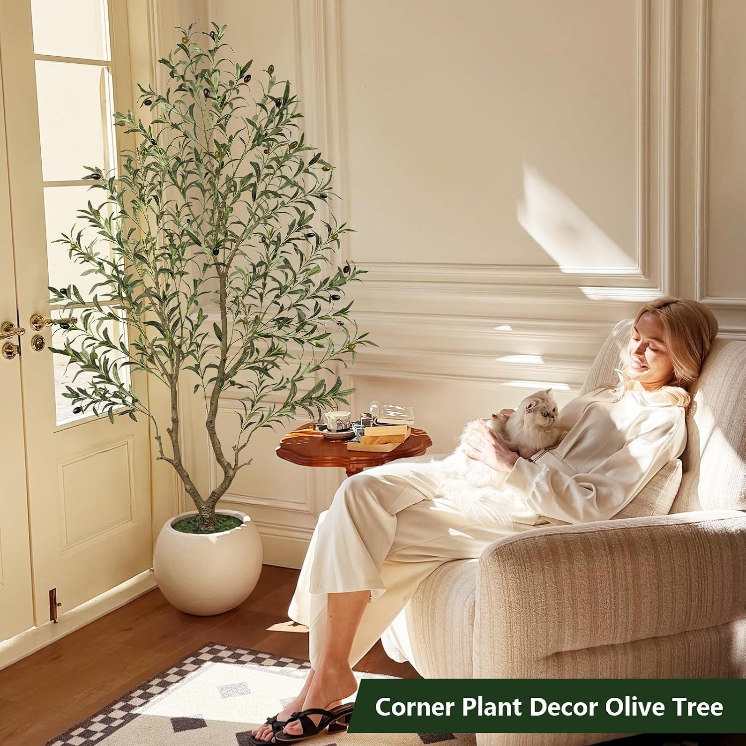 Artificial olive tree in a white round planter placed in a living room corner beside an armchair with a seated woman