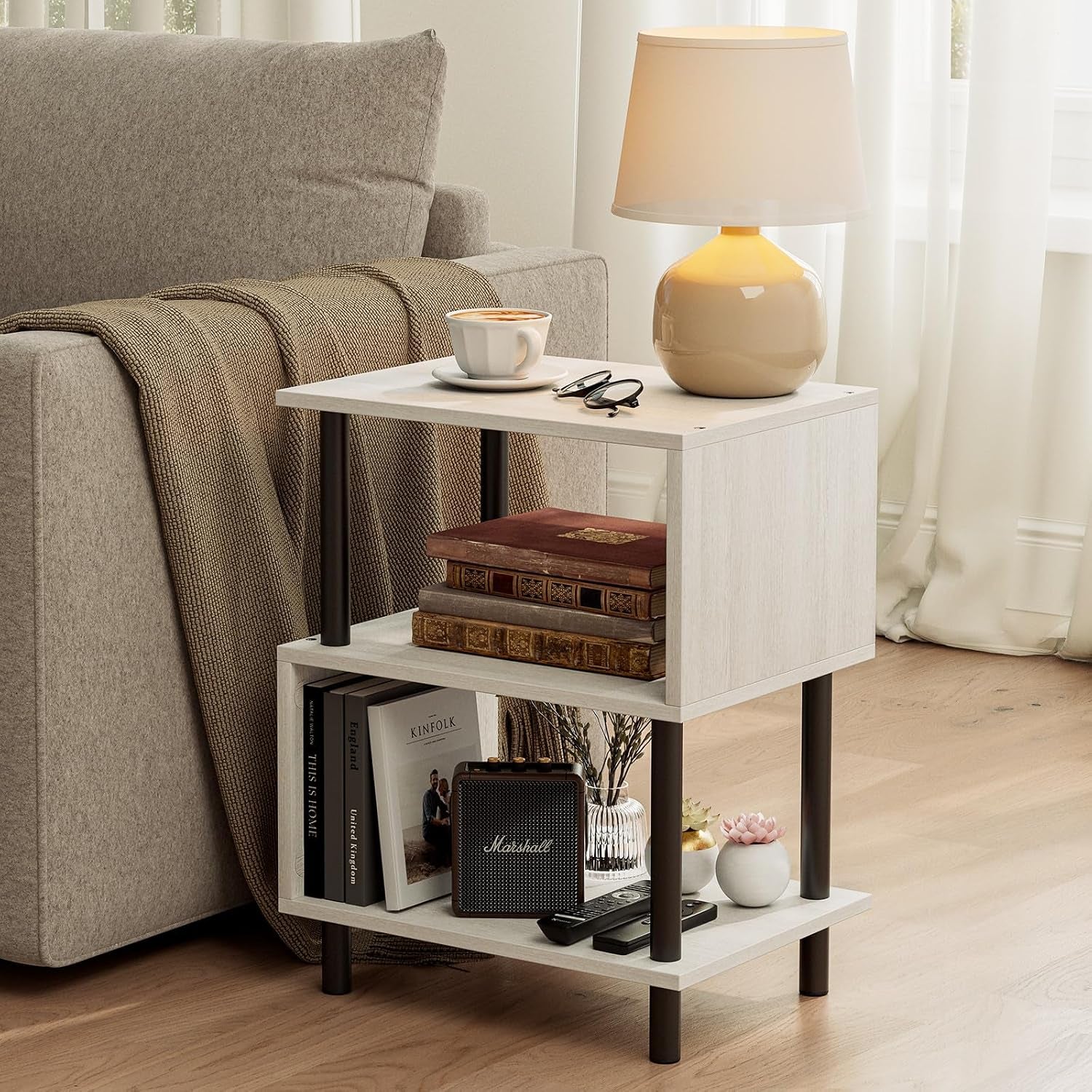 Light wood three-tier side table beside a sofa holding a lamp, books and small speaker