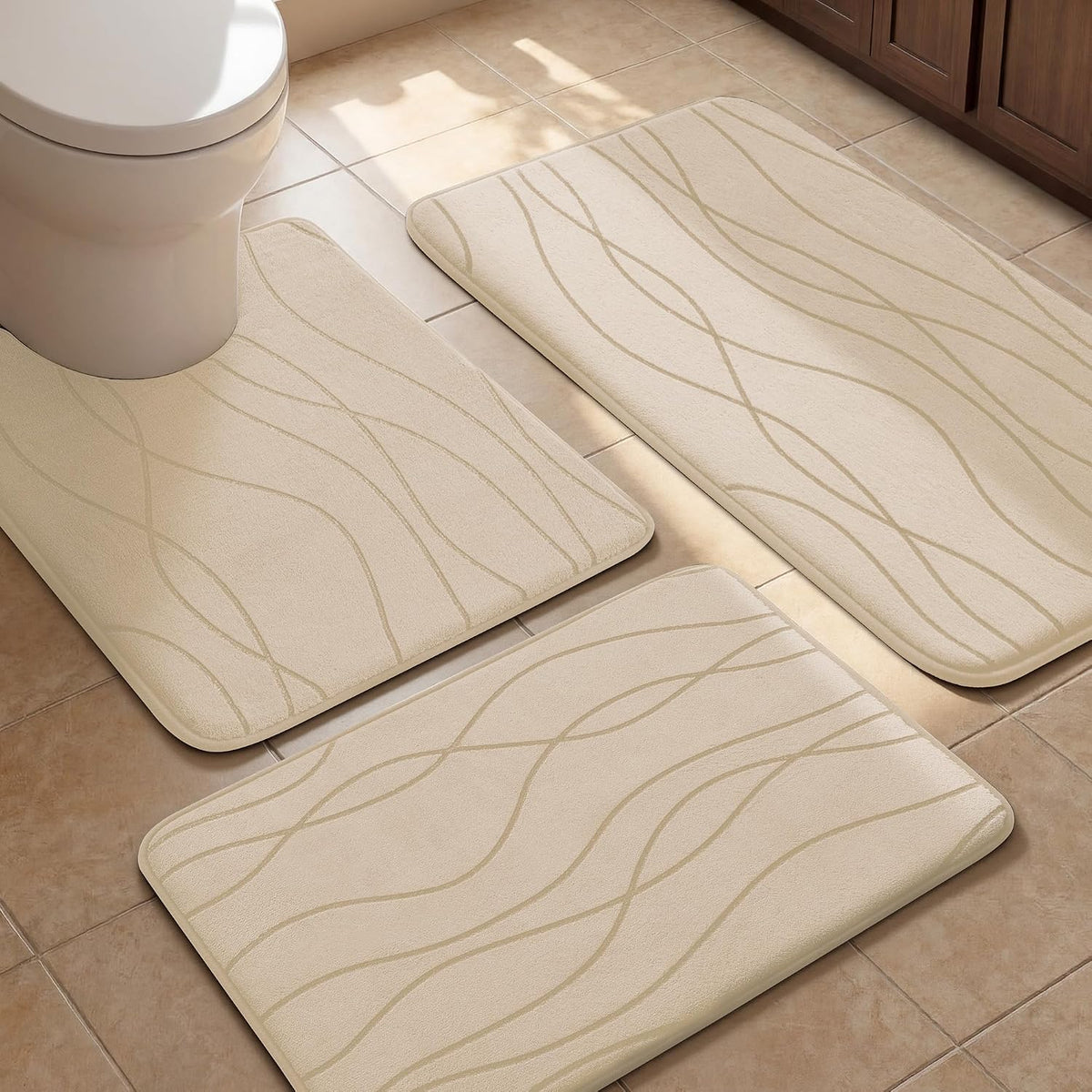 Three beige patterned bath rugs placed on tile around a toilet and wood vanity.