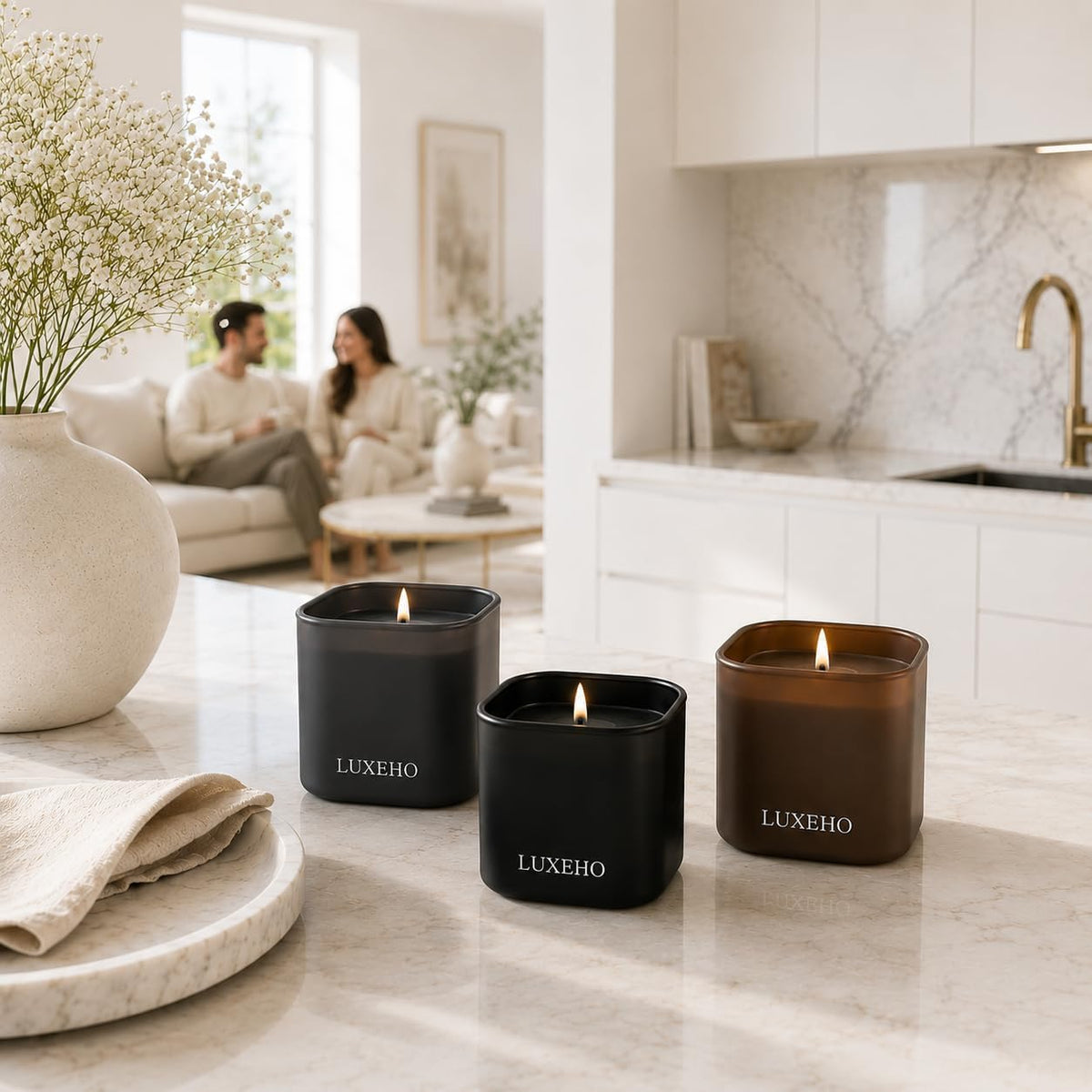 Three lit square candles in a row on a kitchen island with a large vase of white flowers and a blurred couple on a sofa.