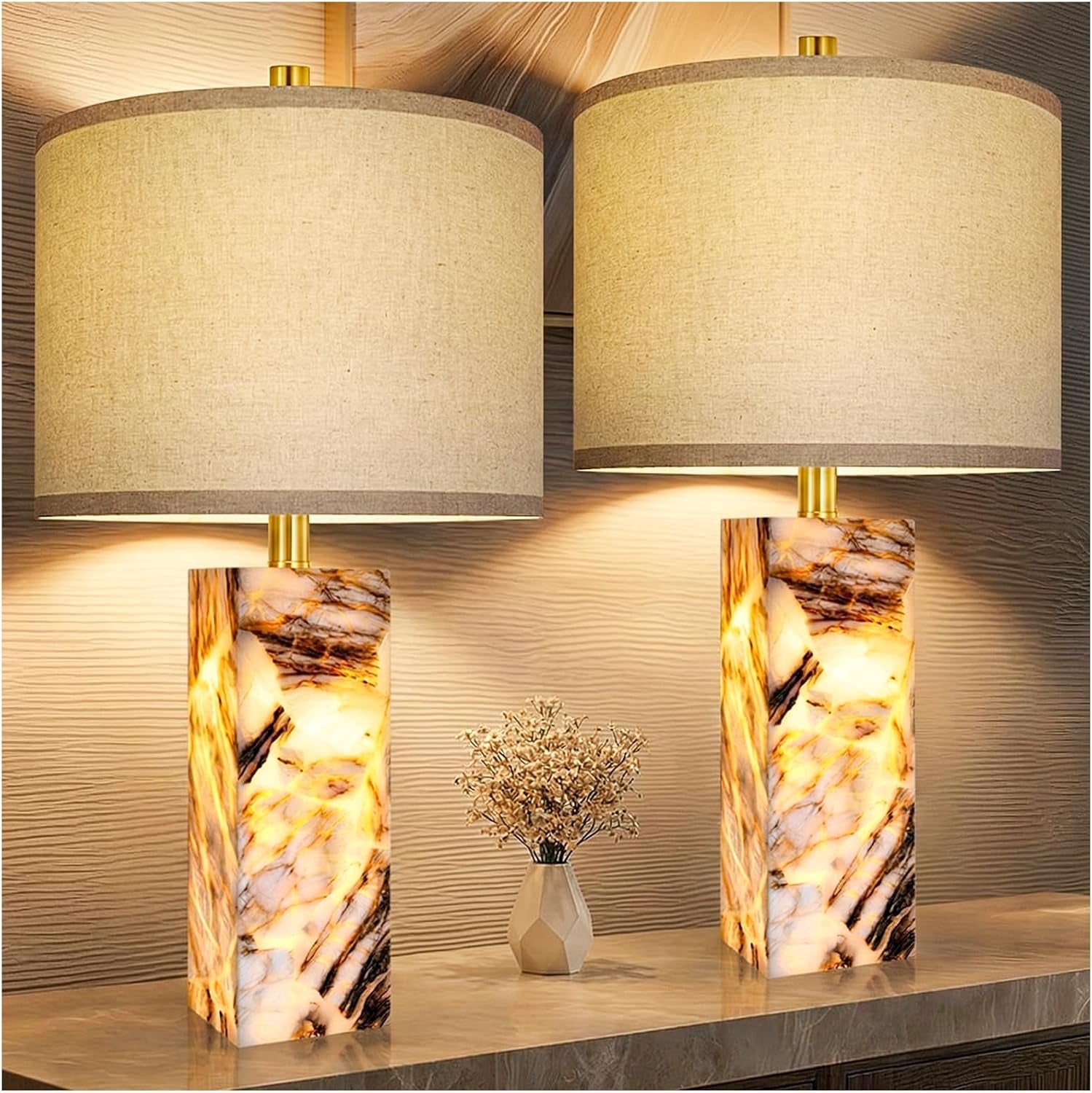 Two glowing alabaster column lamps on a textured wall background with a small geometric vase centered below.