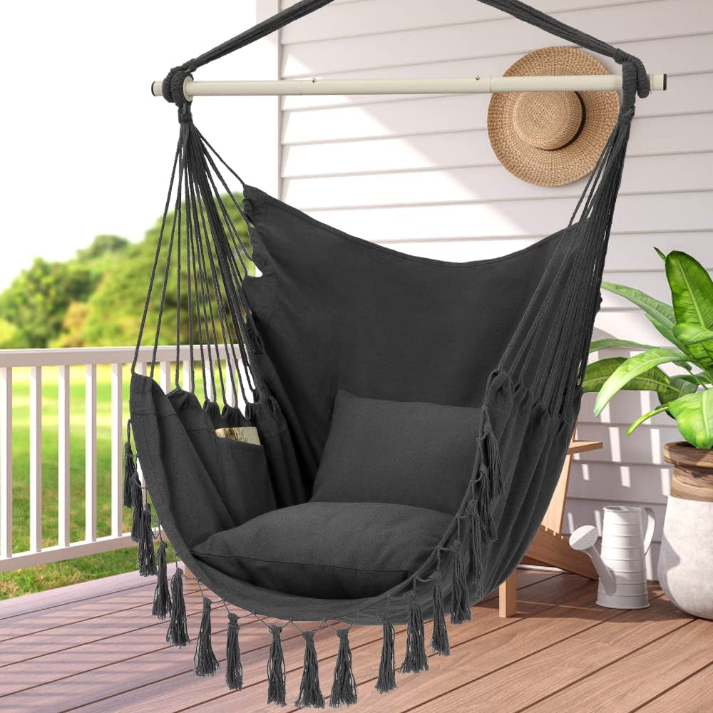 Dark gray hammock chair with cushions hanging from a white bar on a porch, straw hat on the wall behind it.