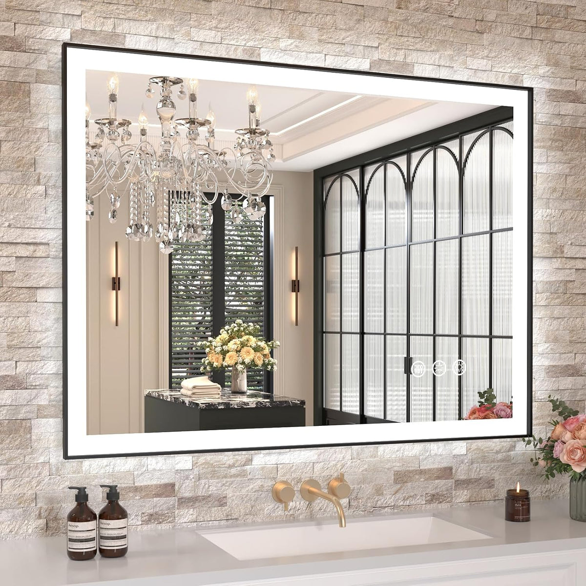 Close view of LED-framed mirror on a stacked-stone wall reflecting chandelier and arched window panels above a single sink