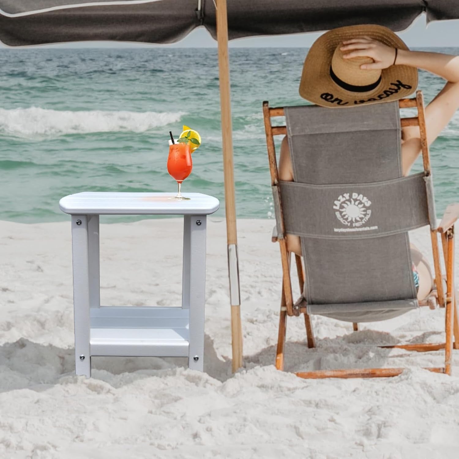 Small white two-tier side table on sandy beach next to a lounge chair and umbrella