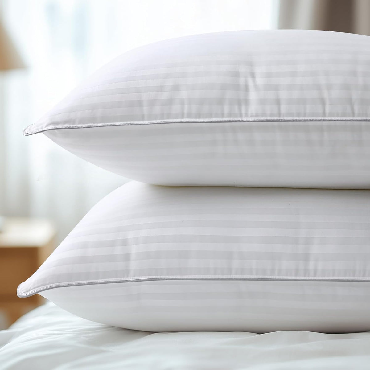 Close side profile of two stacked white pillows on a bed showing subtle horizontal stripe fabric and piped edges.