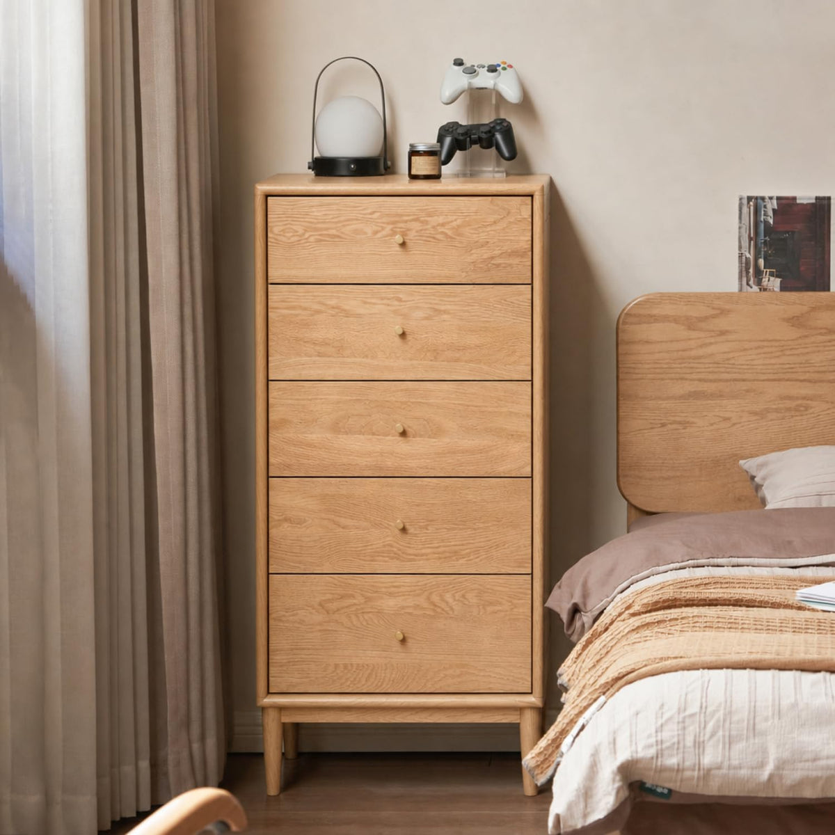 Five-drawer oak dresser styled beside a bed with lamp, controllers and neutral bedding in a softly lit bedroom.