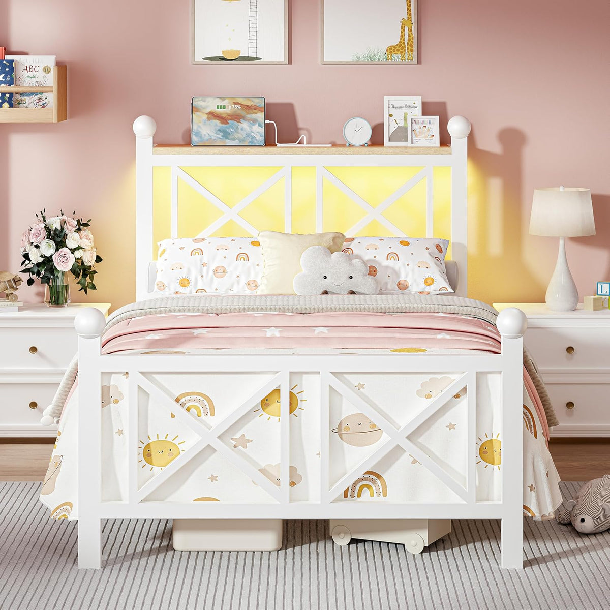 Front view of a white child's bed with sun and rainbow bedding and soft yellow backlighting in a pink room.