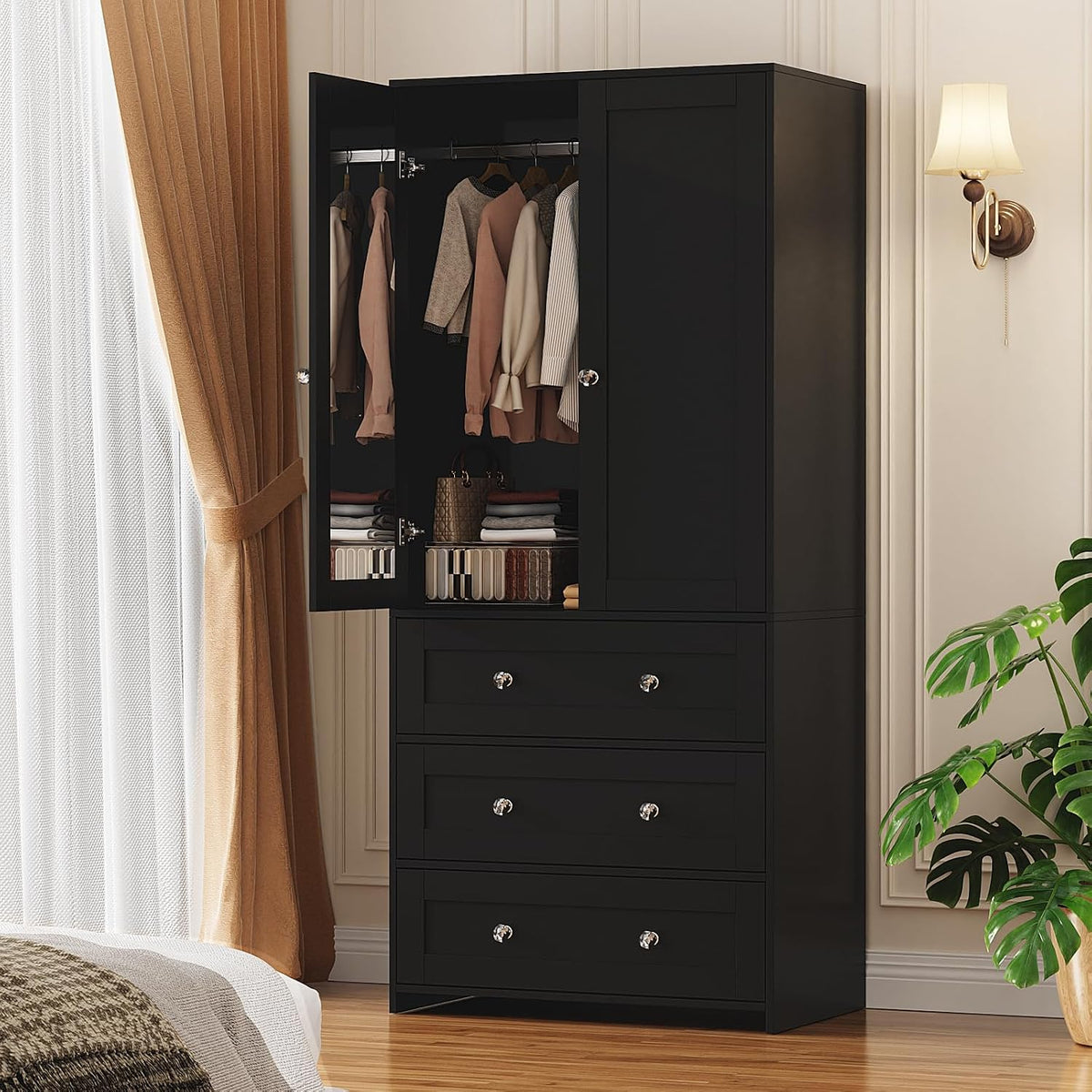 Black armoire wardrobe angled front view with left door open showing hanging clothes and three drawers in a bedroom