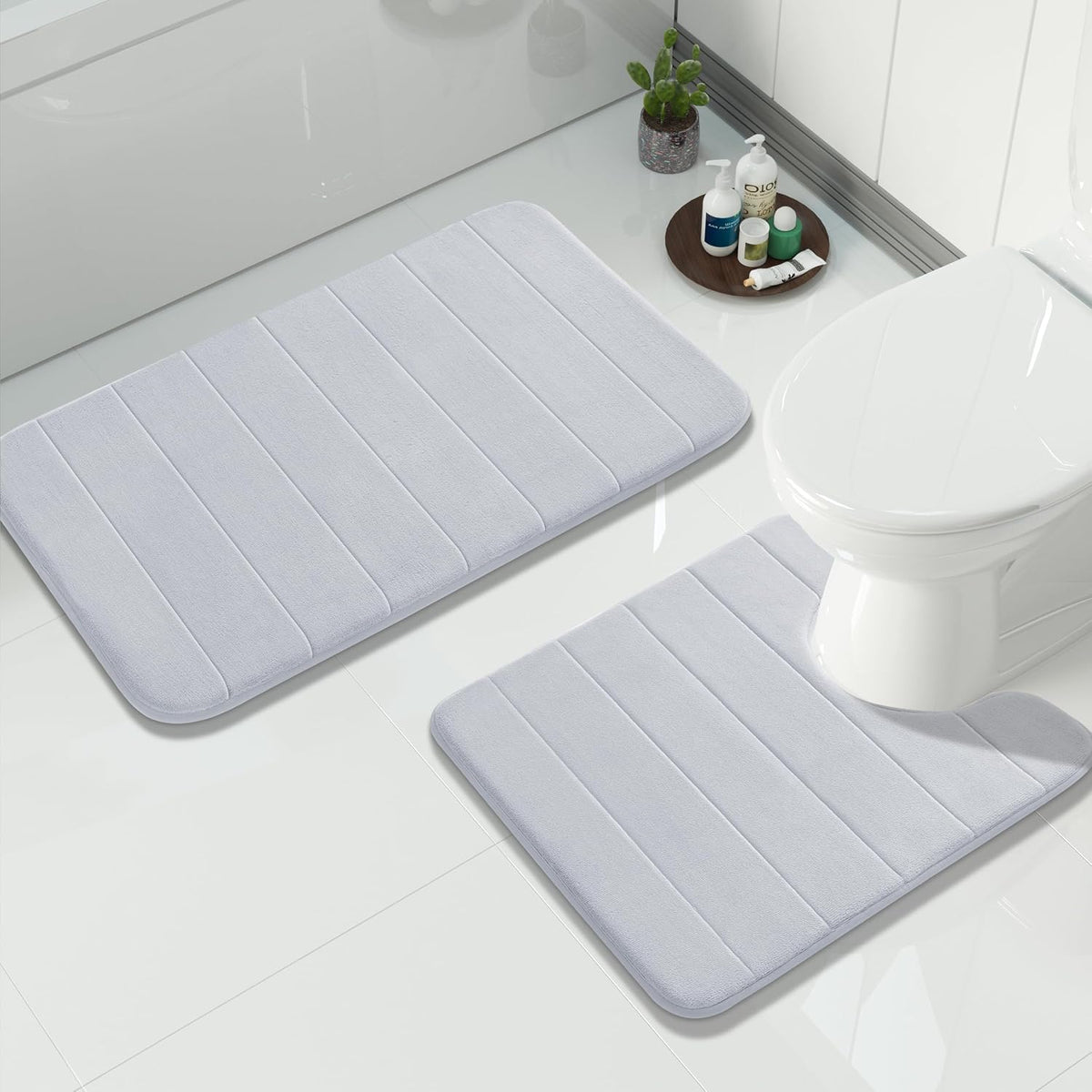 Light gray bath mat and matching U-shaped toilet rug on glossy white tile beside a toilet and small tray of bath items.