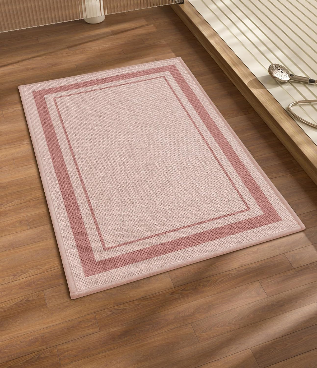 Angled view of a pink rectangular bath rug with bordered design on wood flooring beside a raised tub.