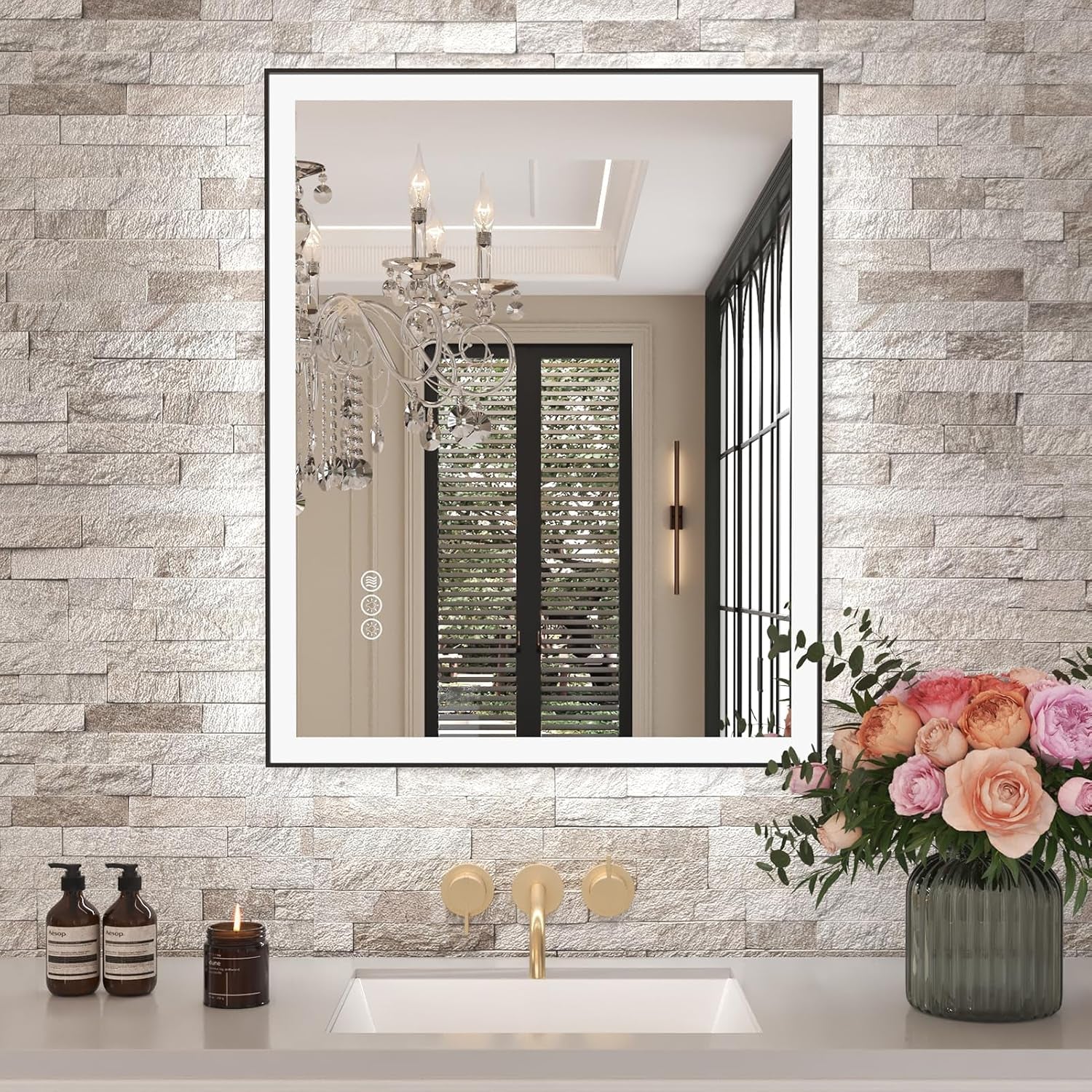 Rectangular LED-lit mirror on a light stone wall above a sink with gold faucet and a vase of roses.