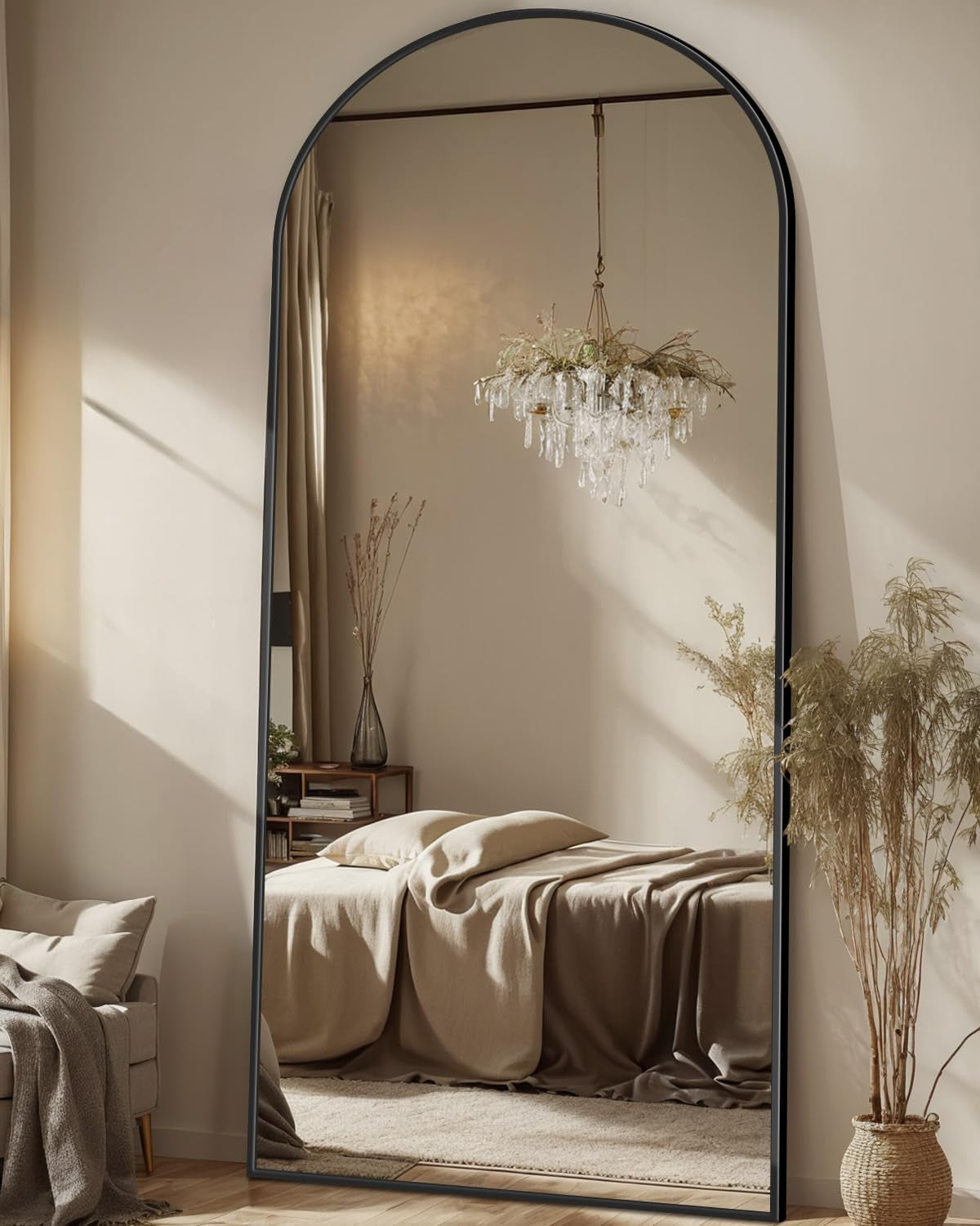 Arched full-length mirror with thin black frame reflecting a bed and chandelier in a softly lit bedroom.