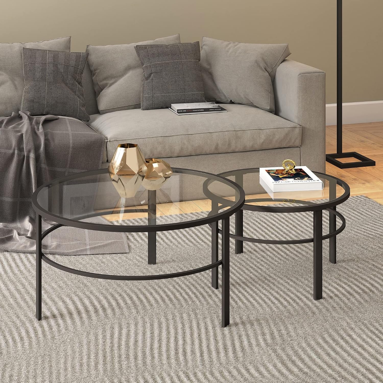 Two round glass-top tables in front of a gray sofa, decorated with two gold geometric vases and a book on a textured rug.