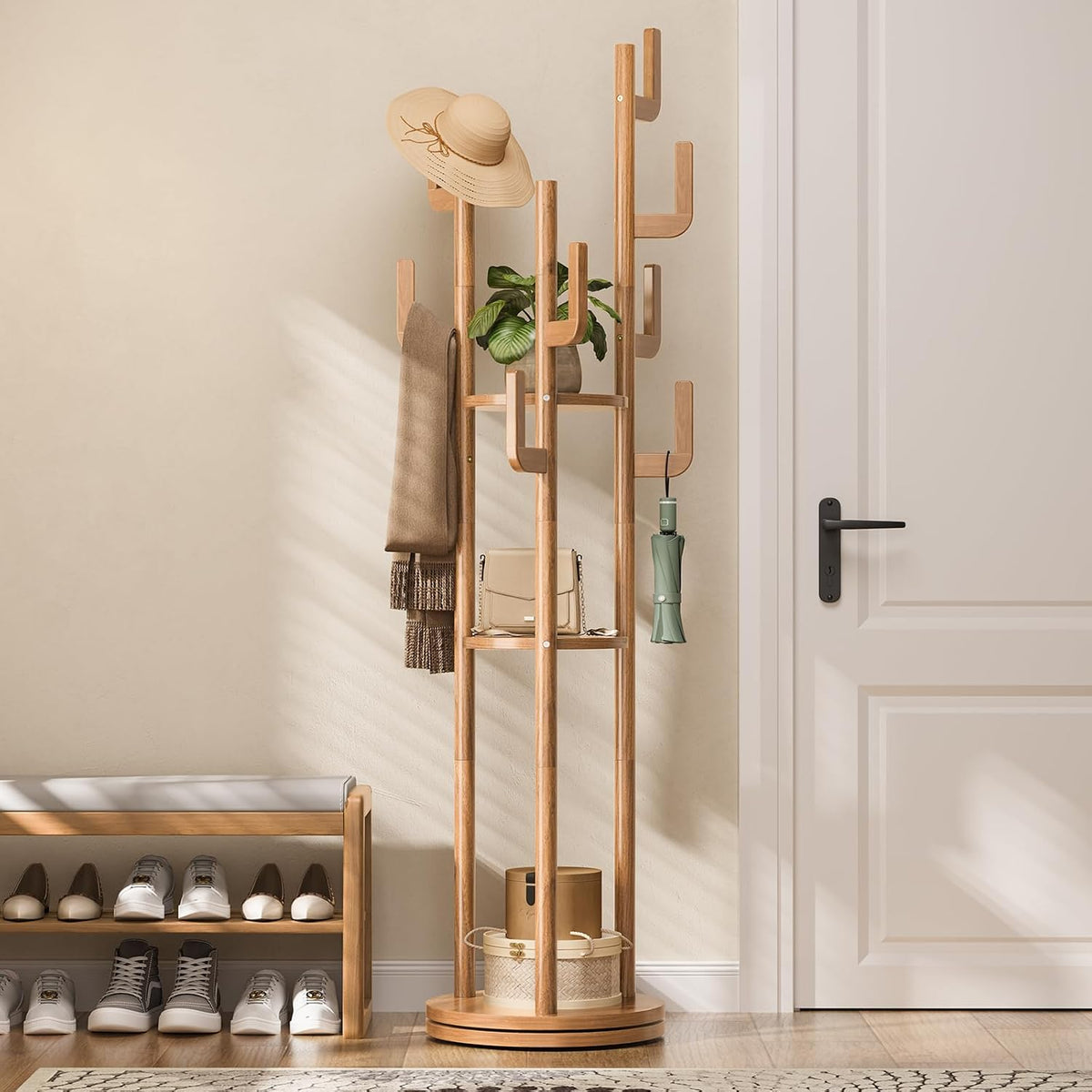 Entryway view of a light-wood rotating coat rack beside a closed white door, holding a hat, scarf, plant, bag and umbrella.
