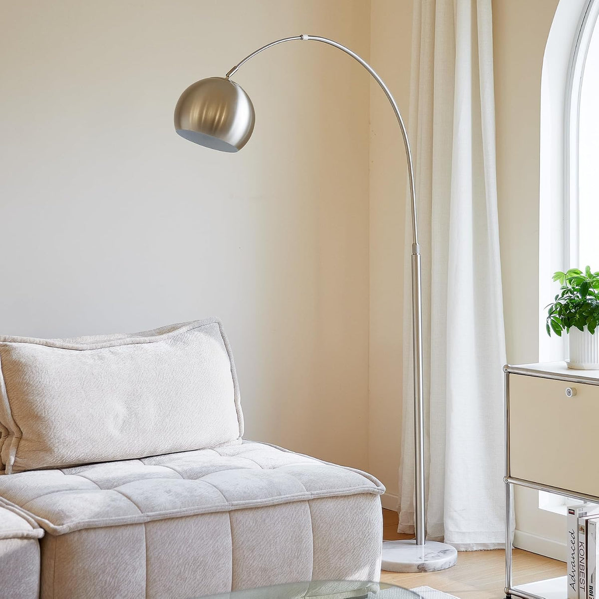 Brushed silver arc lamp next to a light chaise and a window with sheer curtains, marble base visible