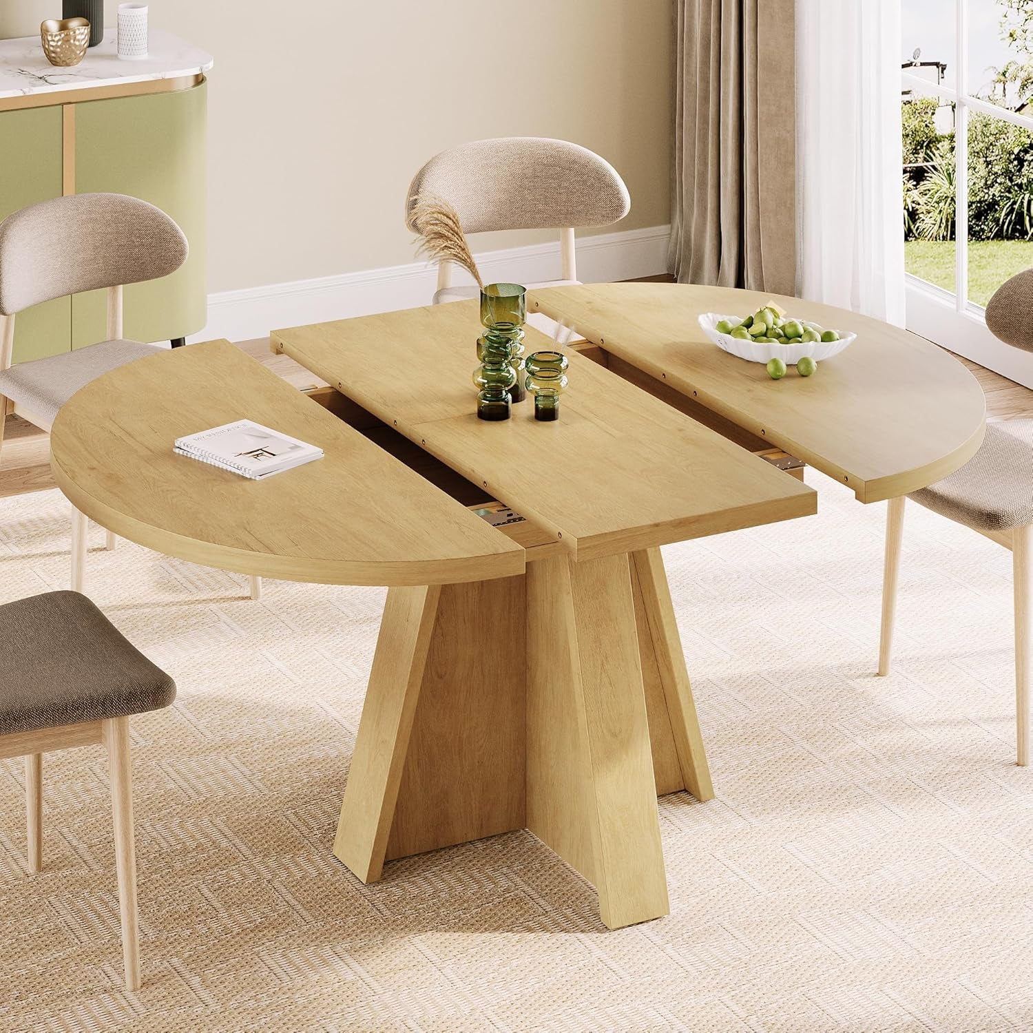 Light wood round dining table shown extended with center leaf, four upholstered chairs around.