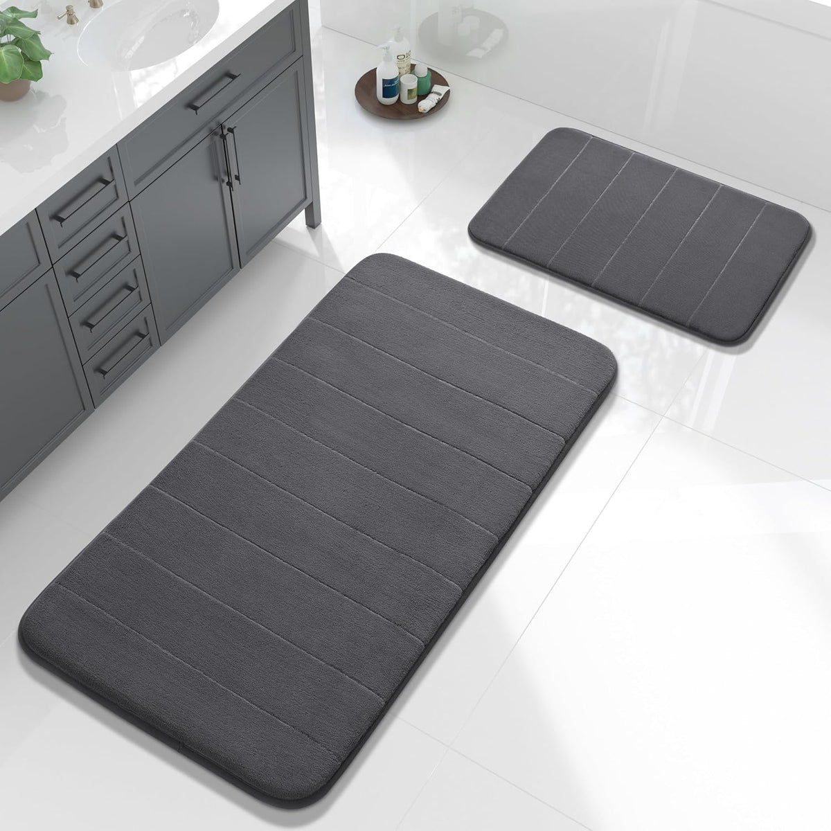 Large rectangular and smaller rectangular dark gray bath mats placed on a tiled bathroom floor near a gray vanity.