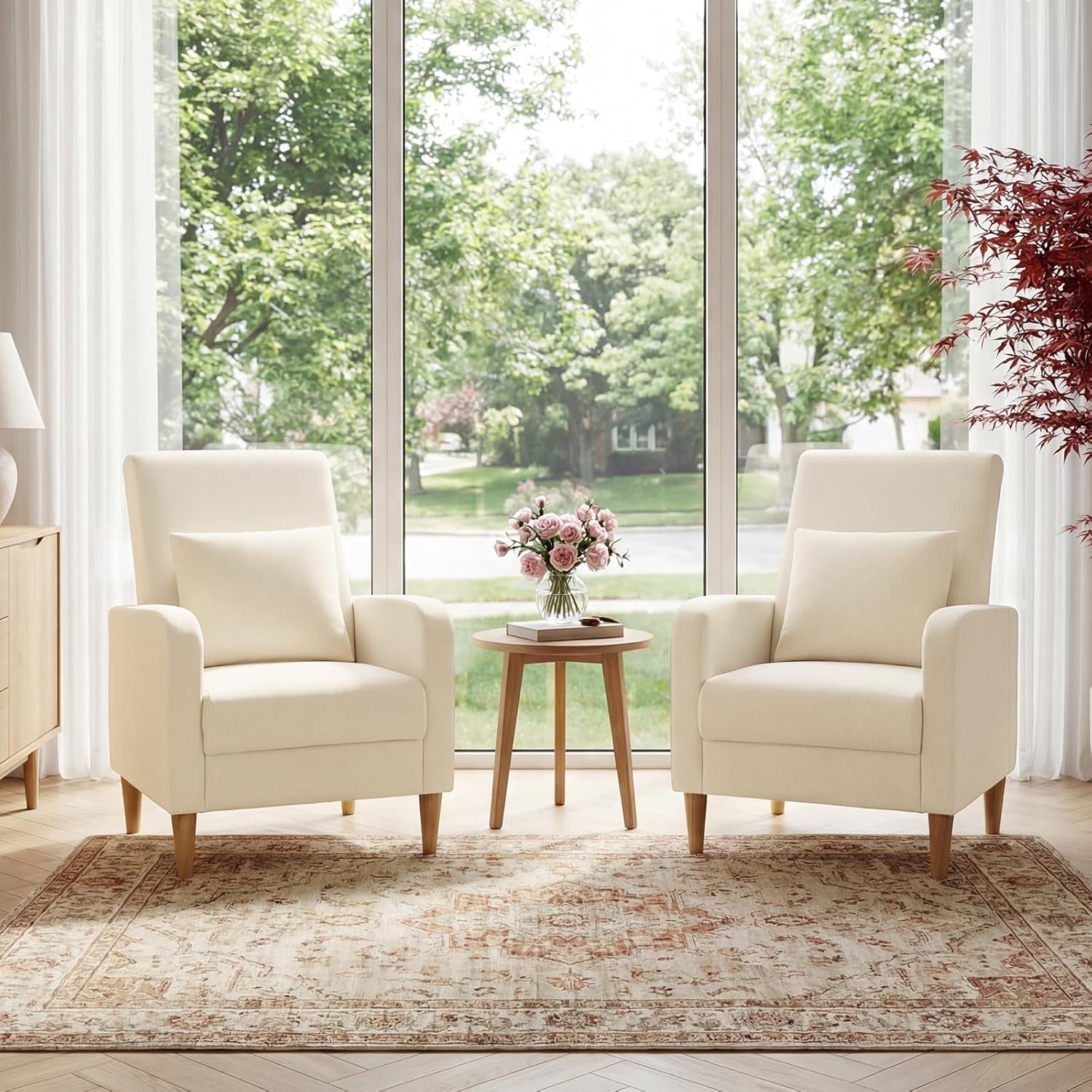 Two beige fabric accent chairs with wooden legs arranged around a small round table by a large window
